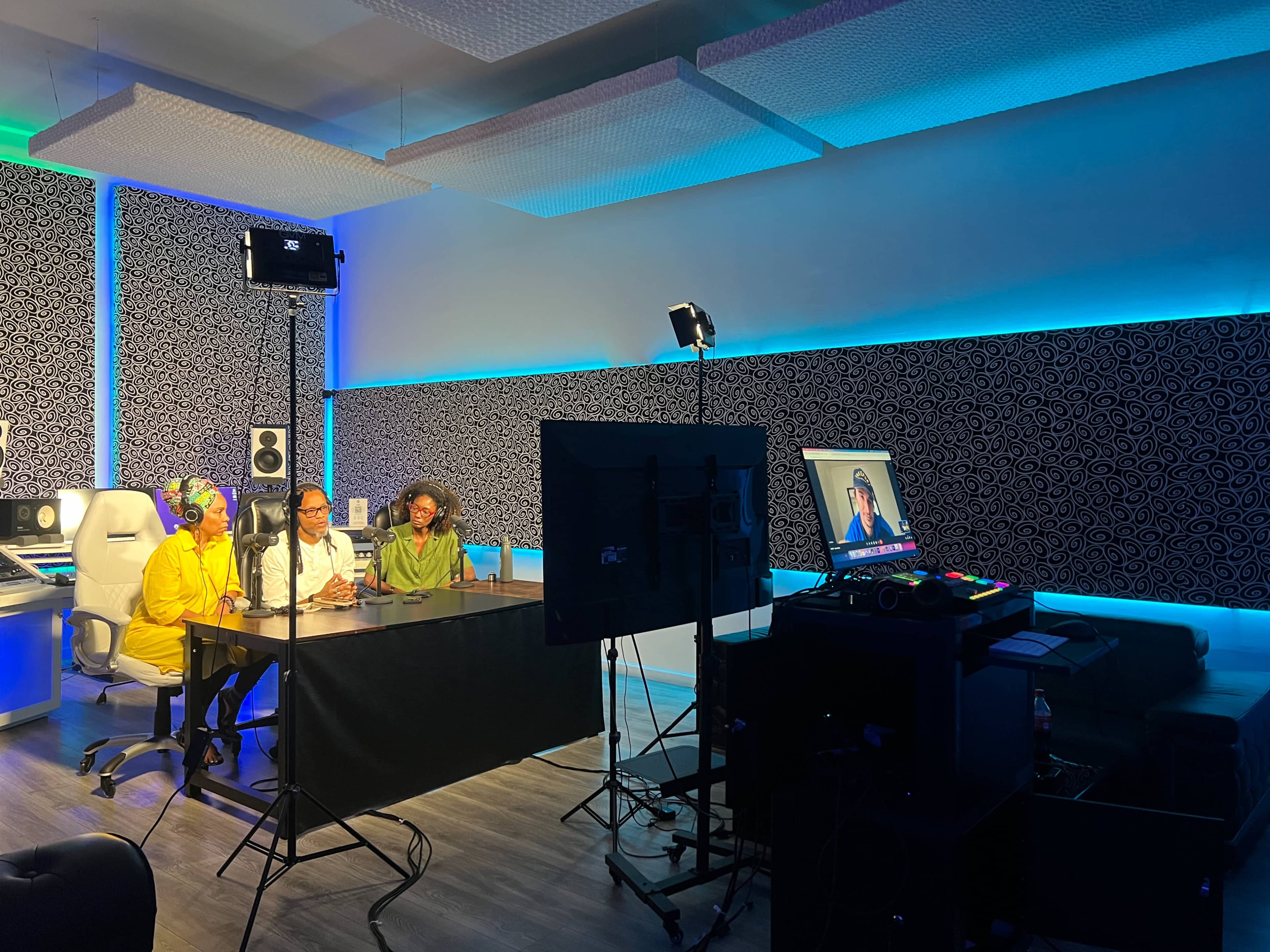 A group of three people sits at a table in a studio, facing a monitor displaying another individual, with studio equipment and colorful lighting in the background.