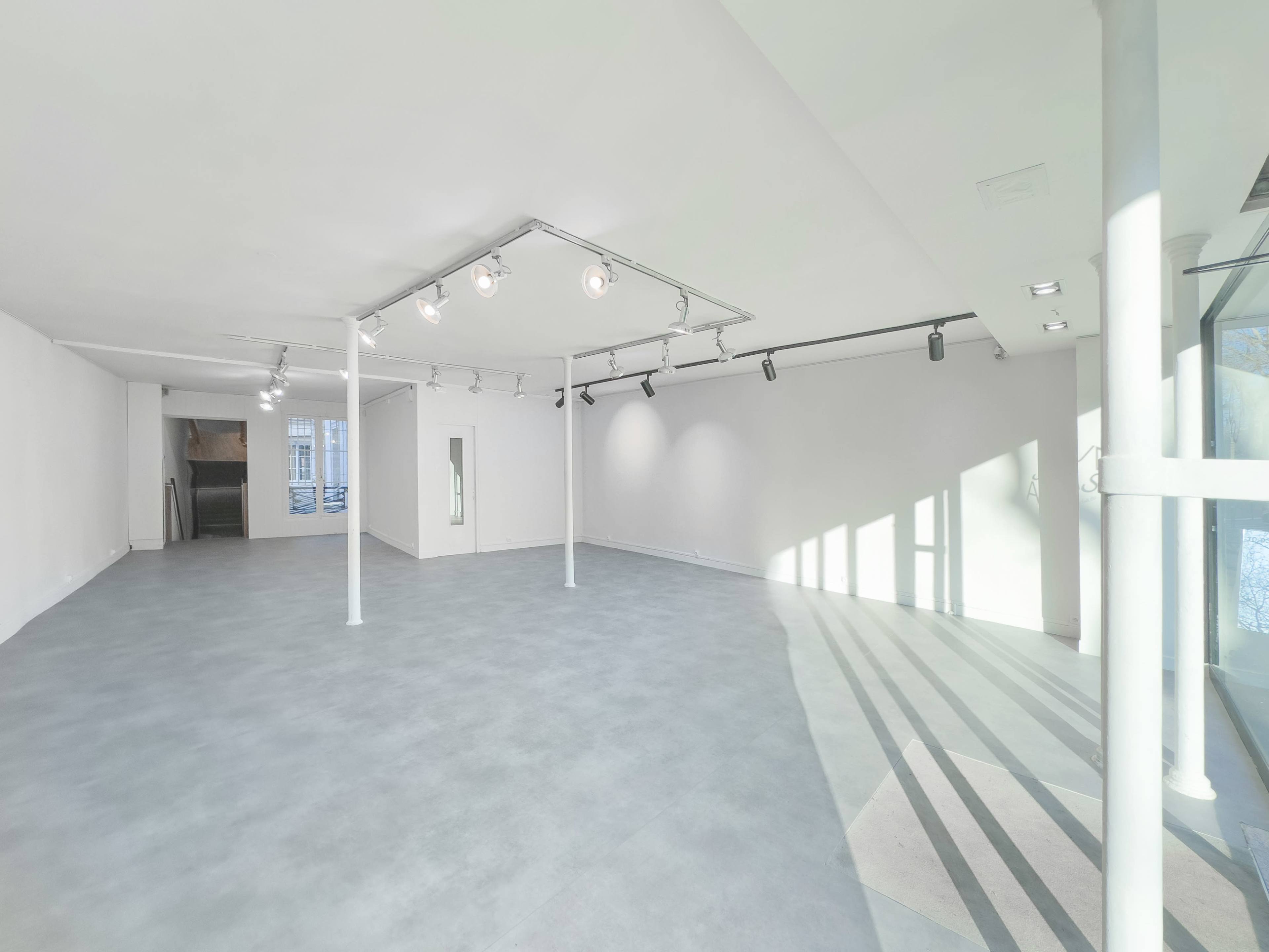 The image shows an empty, well-lit gallery space with white walls and a light gray floor.