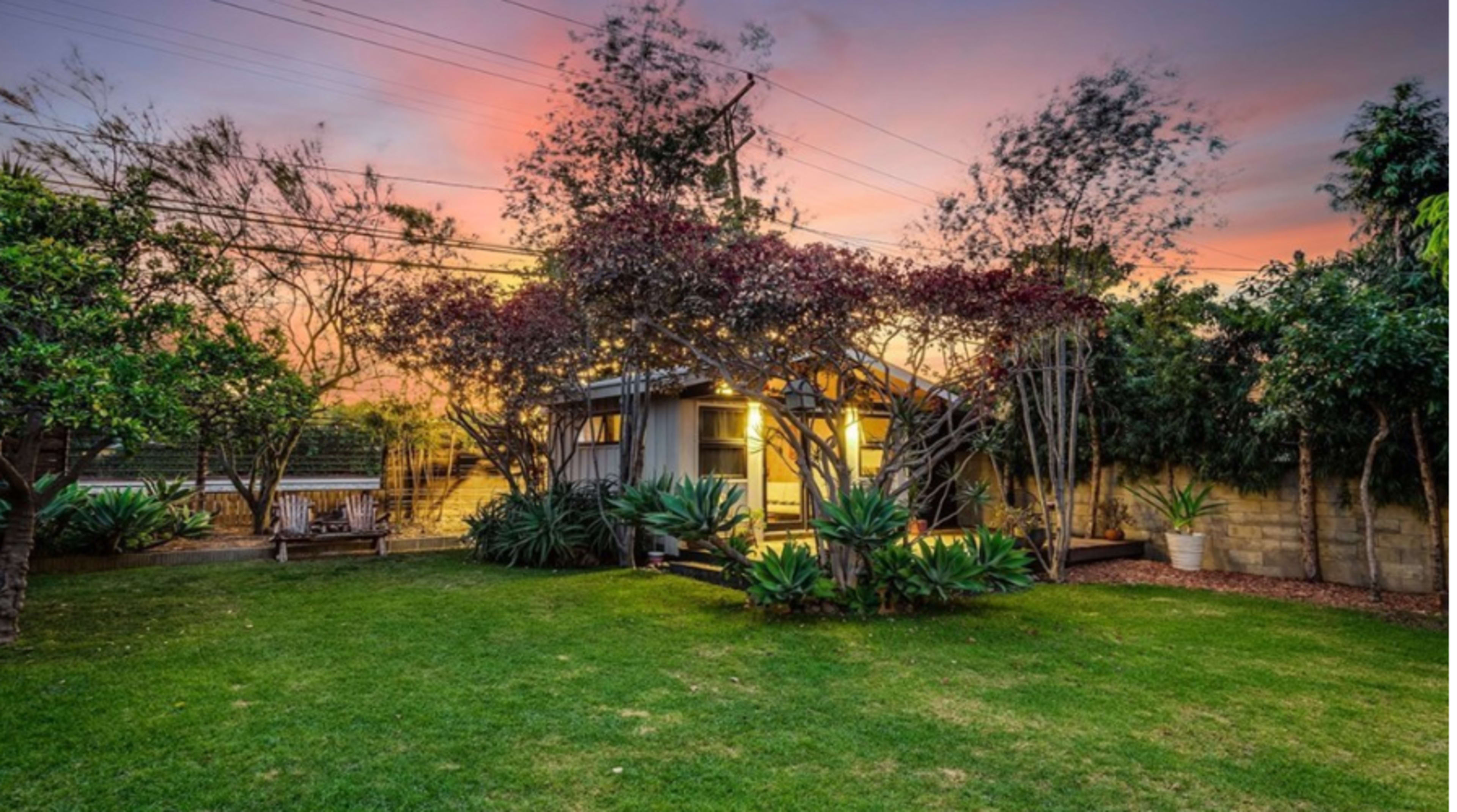 A house with a well-maintained lawn and surrounding trees is illuminated by evening light under a colorful sunset sky.