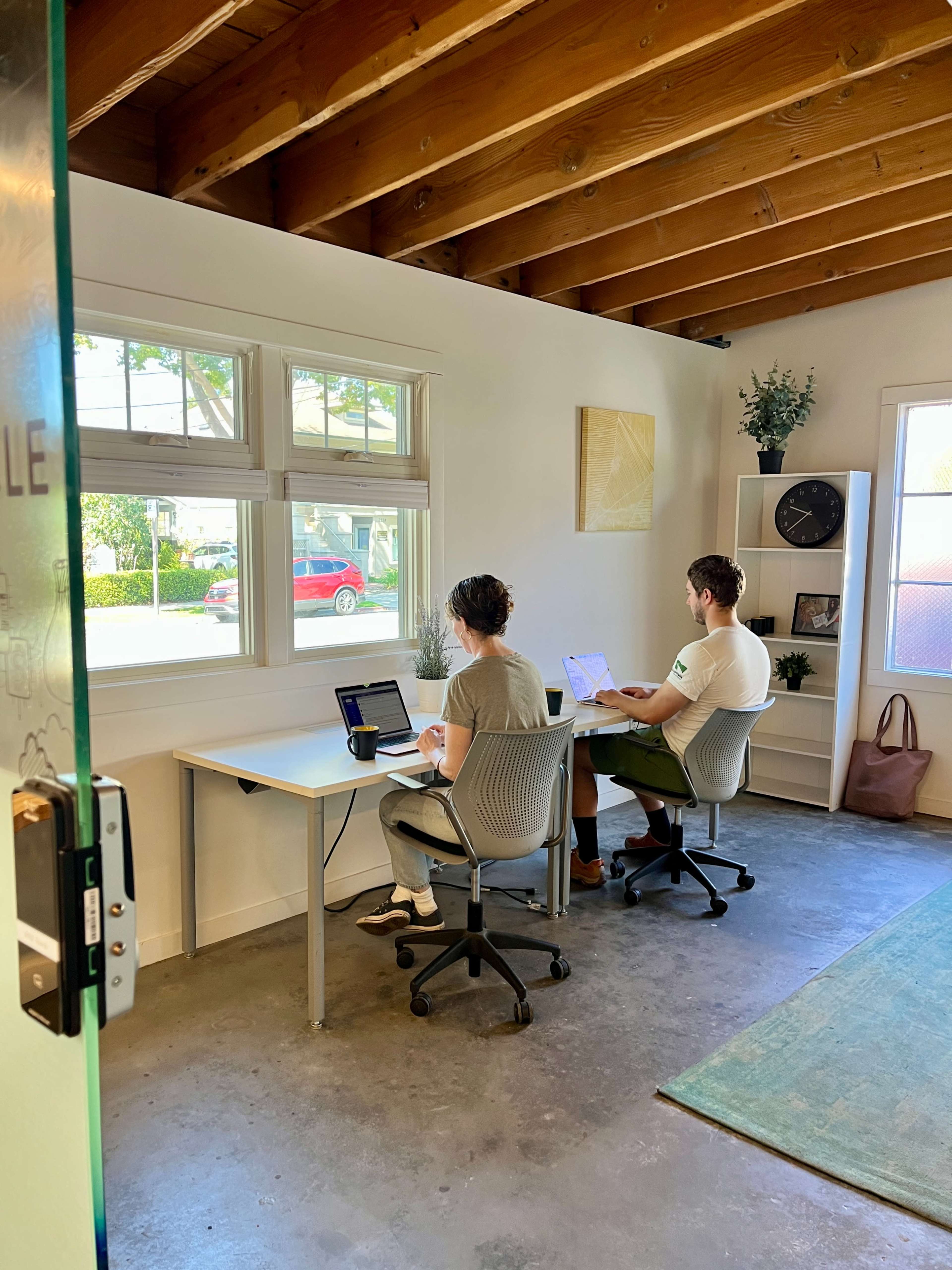 Two people sit at separate desks in a bright room with large windows and a light color scheme, working on laptops.