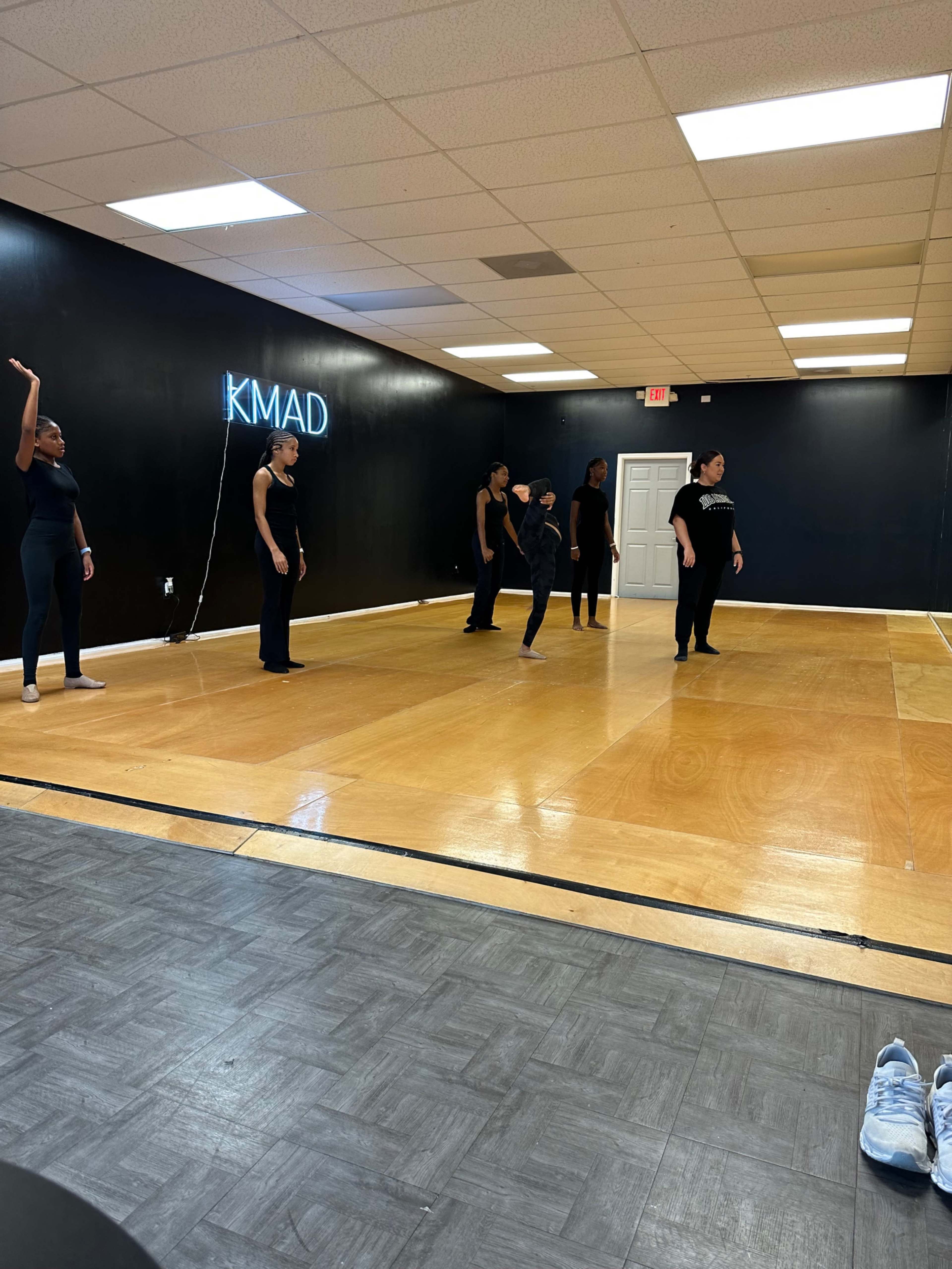 A group of six dancers is positioned in a dance studio with a mirrored wall, wooden floor, and a neon sign that reads "KMAD."