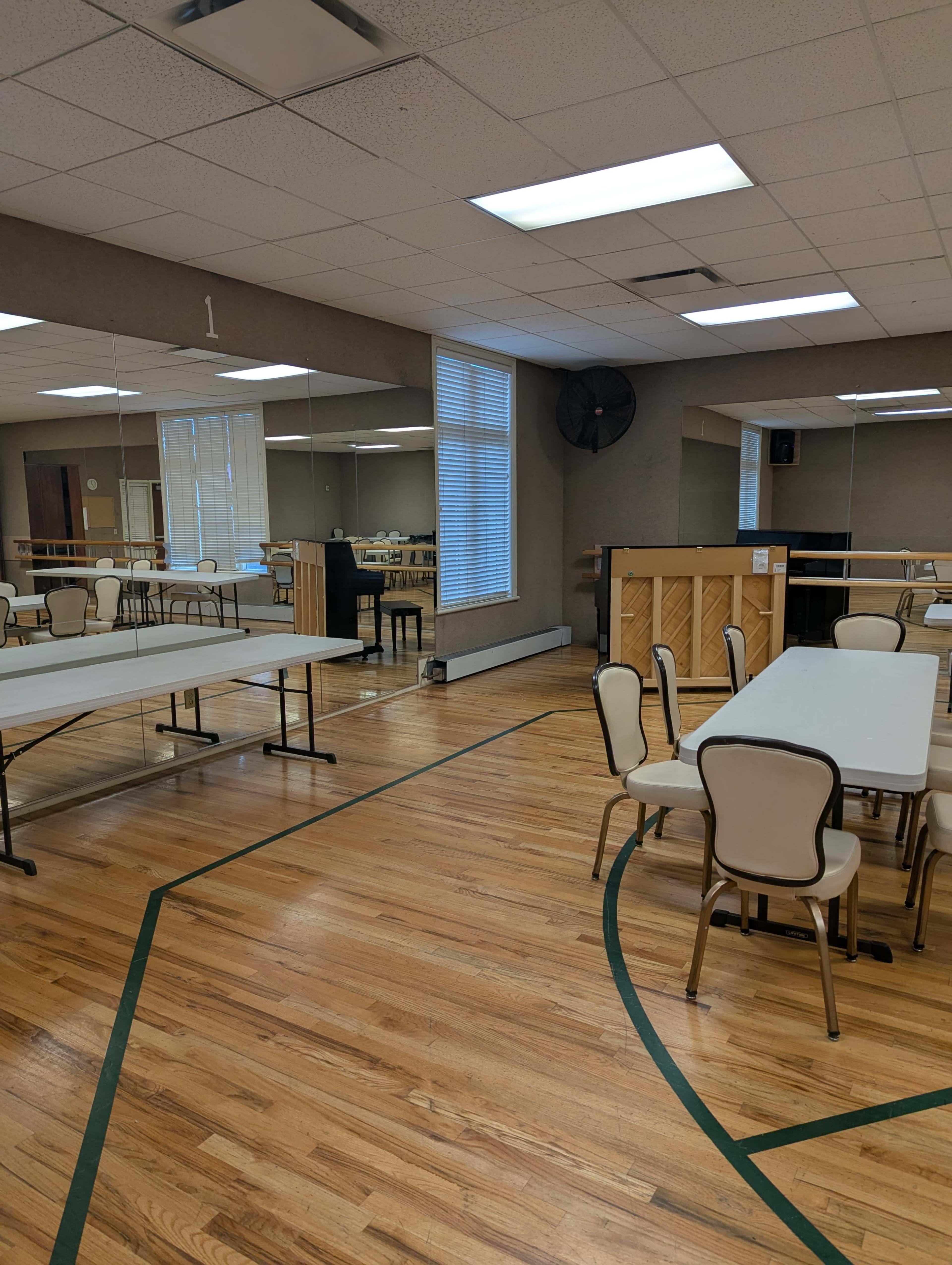 The image shows a spacious room with wooden floors, several tables and chairs set up, a piano against one wall, and large mirrors reflecting the space.