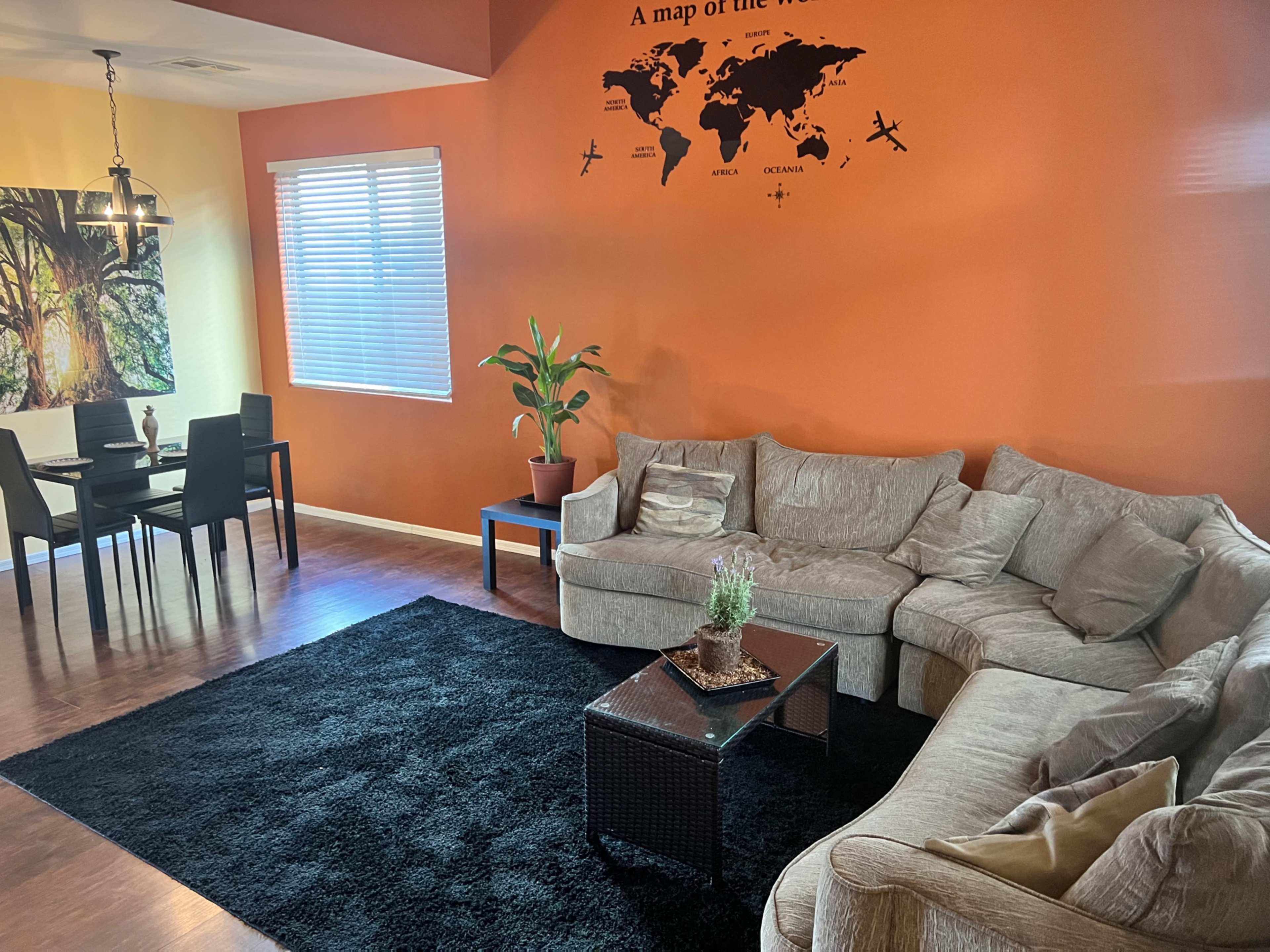 The room features a sectional sofa, a black area rug, a small coffee table, a potted plant, and a dining area with a table and chairs against a brightly colored wall with a world map design.