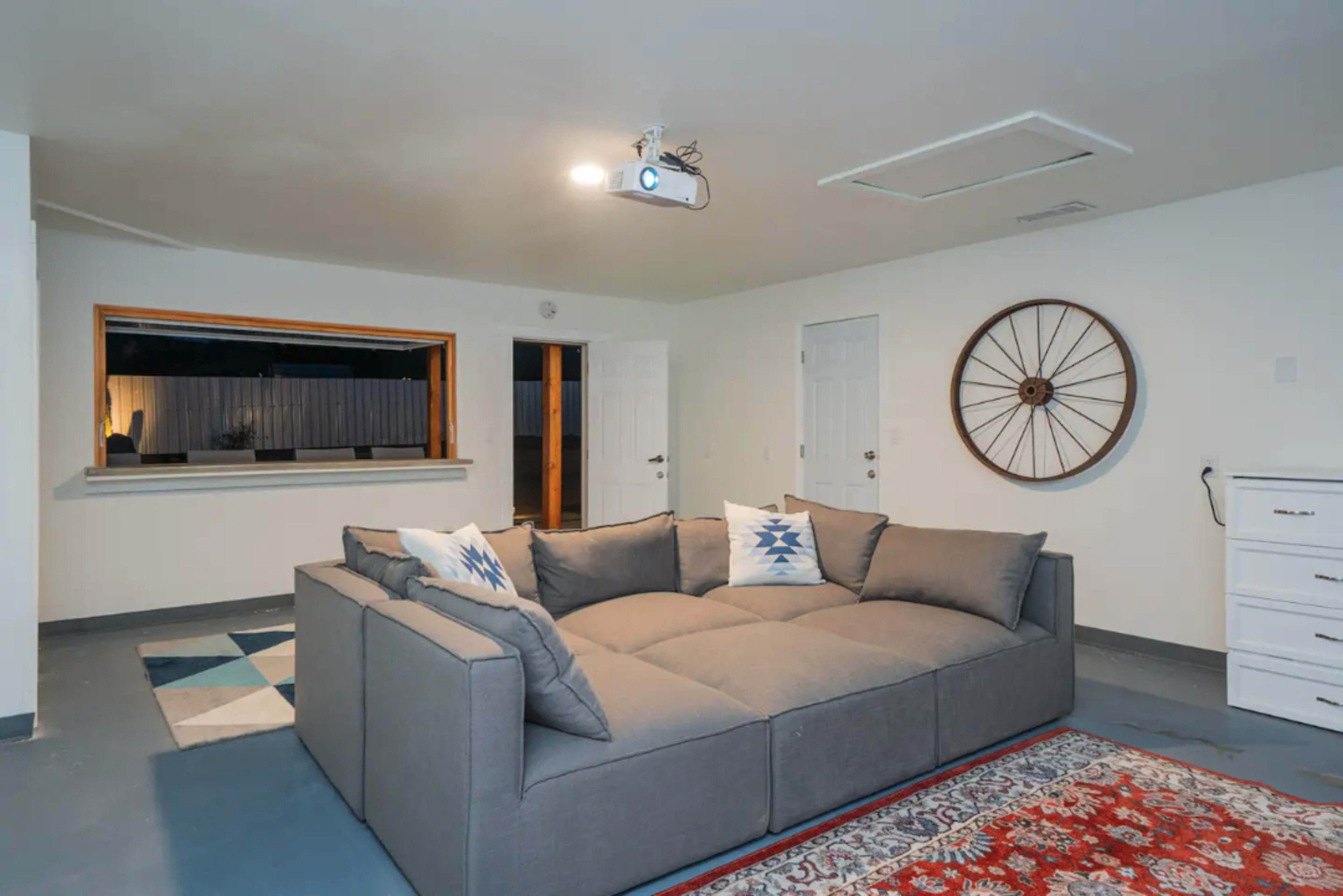 The scene features a gray sectional couch with decorative pillows, a wall-mounted projector, and a large rug in a minimalist living room with a window facing an outdoor space.