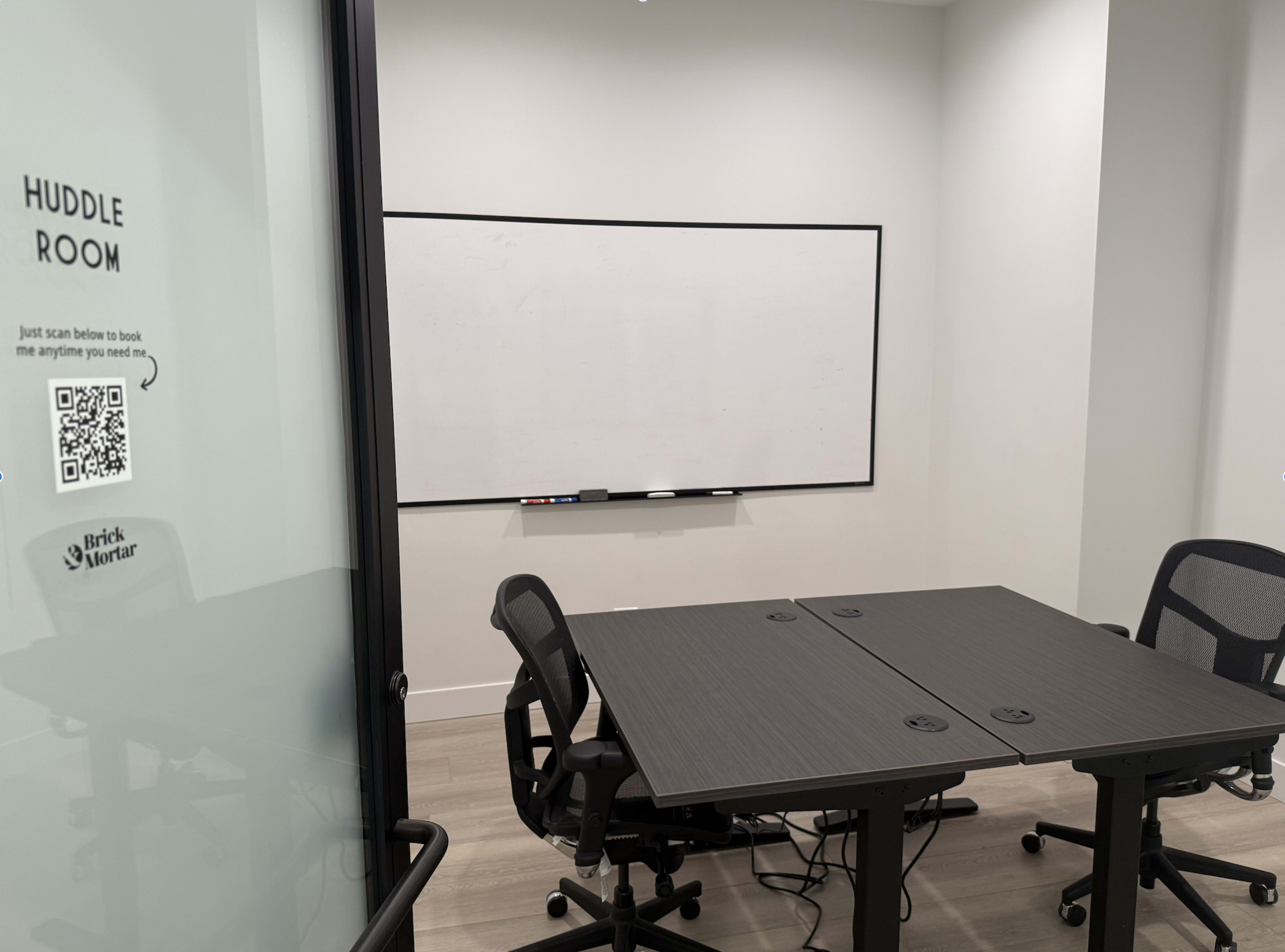 The image shows a small huddle room featuring a table with four chairs and a large whiteboard on the wall, with a QR code on the glass door for booking.