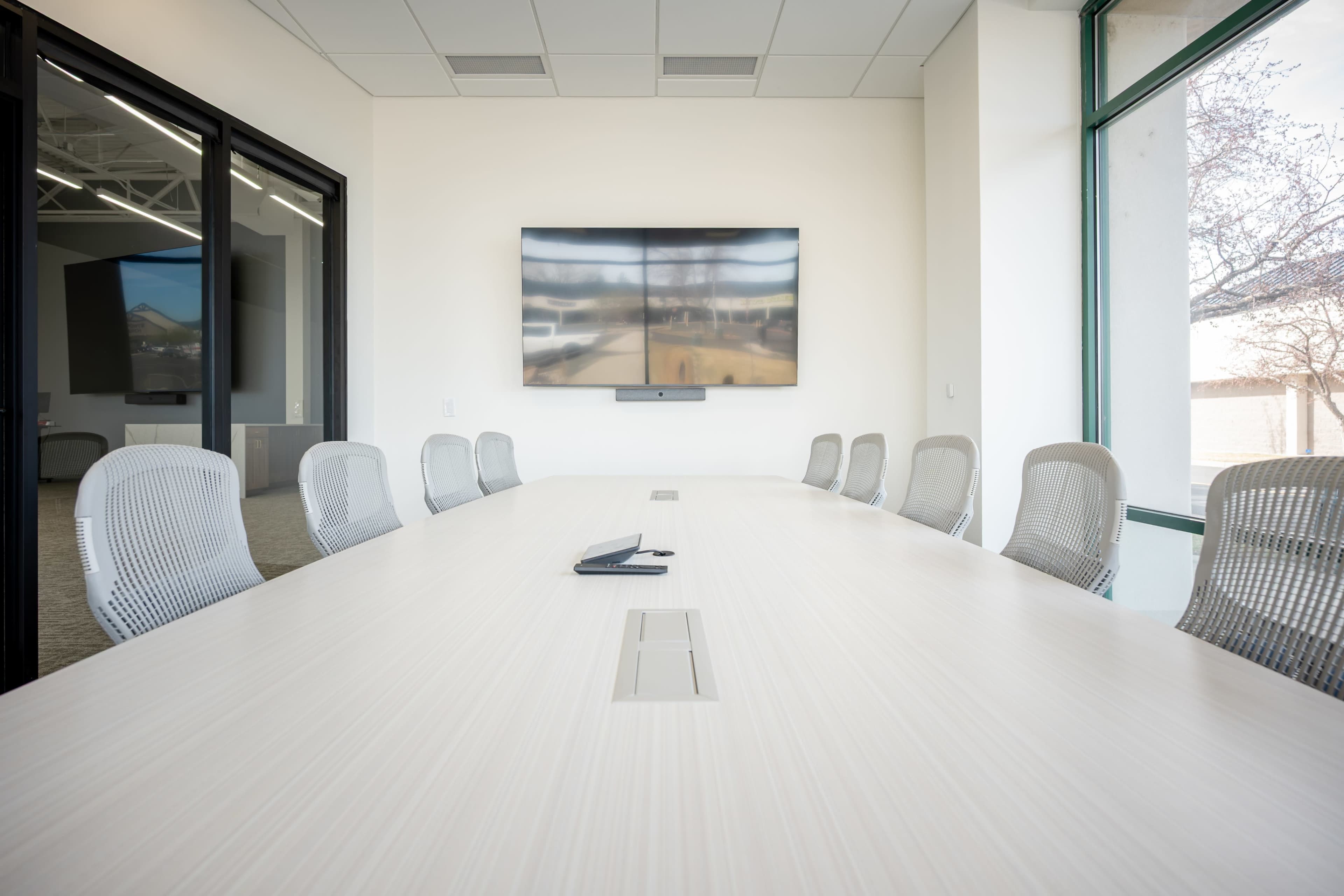 A modern conference room features a long table with multiple chairs and a large flat-screen TV mounted on the wall.