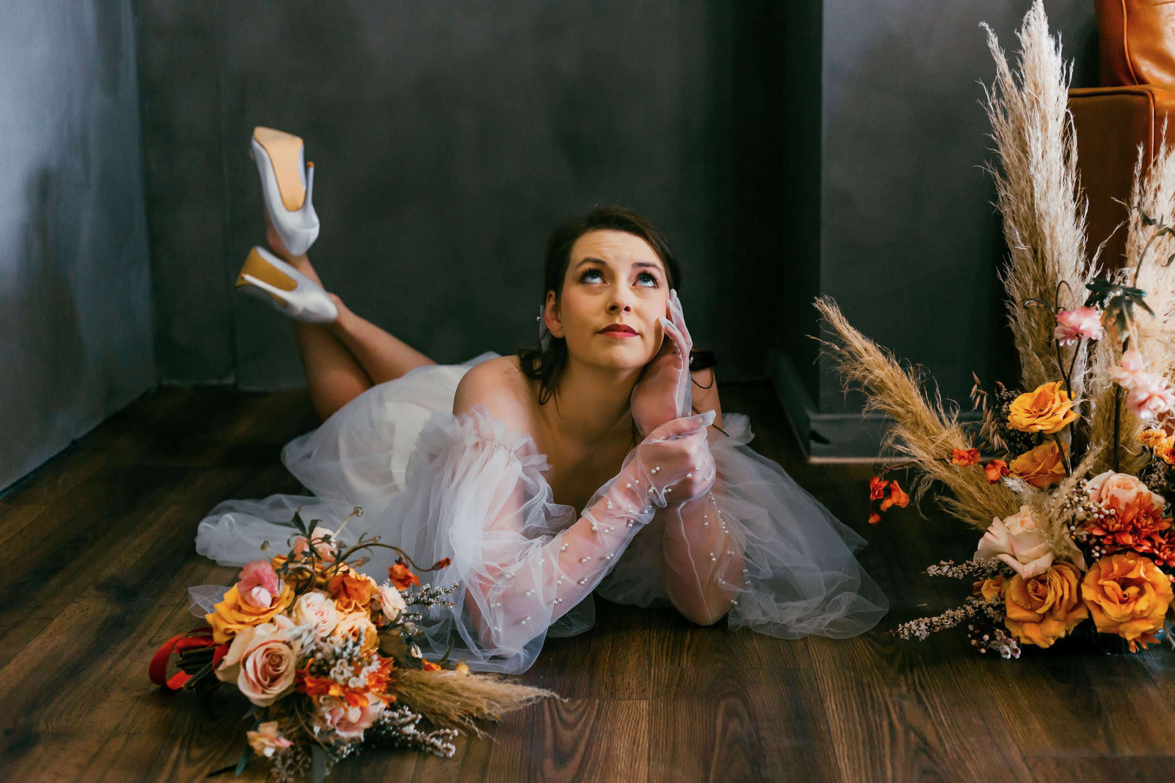 A bride in a white dress lies on a wooden floor, resting her chin on her hand, surrounded by floral arrangements.