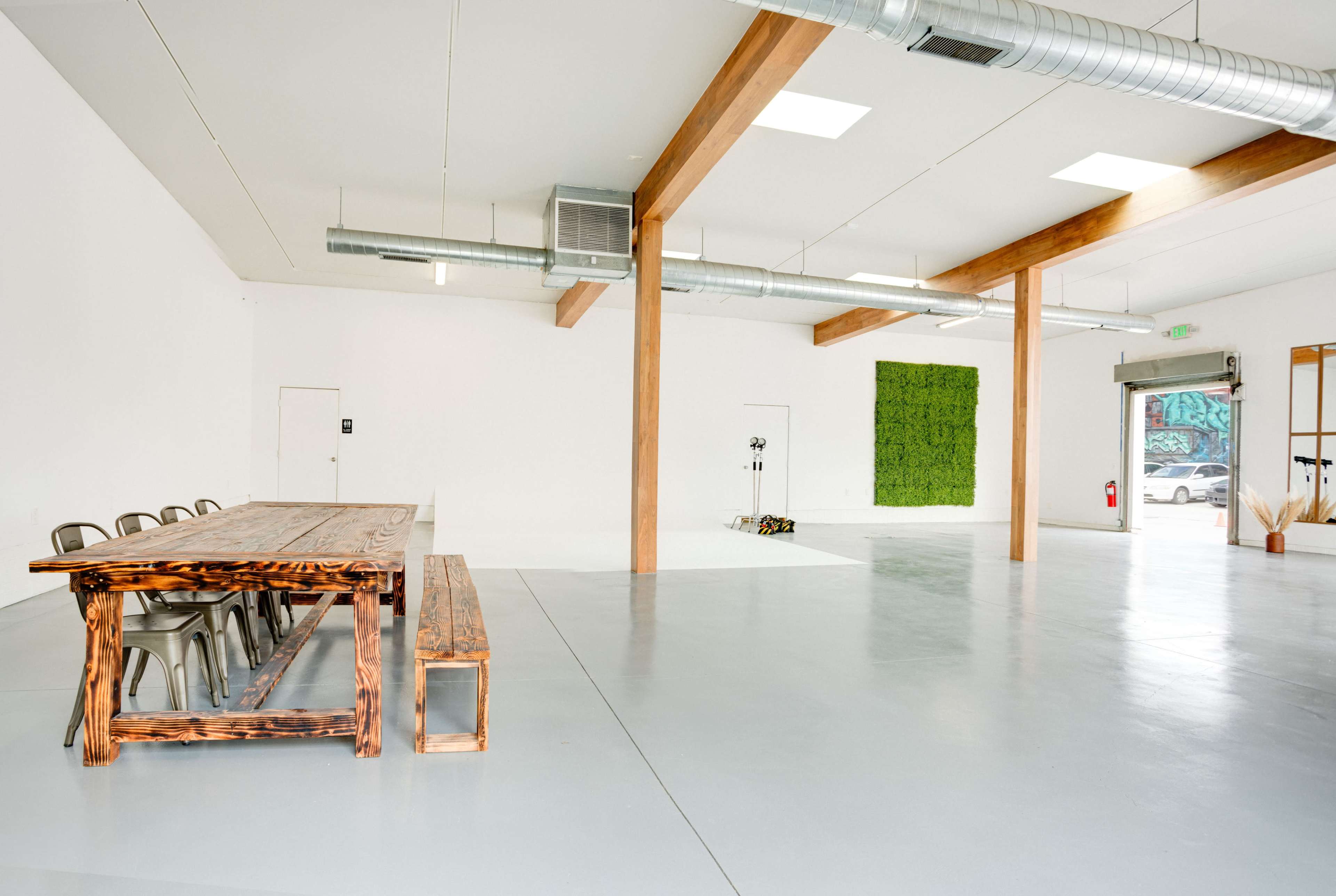 A spacious, empty room with a large wooden table and benches, gray flooring, white walls, and a vertical green wall feature.