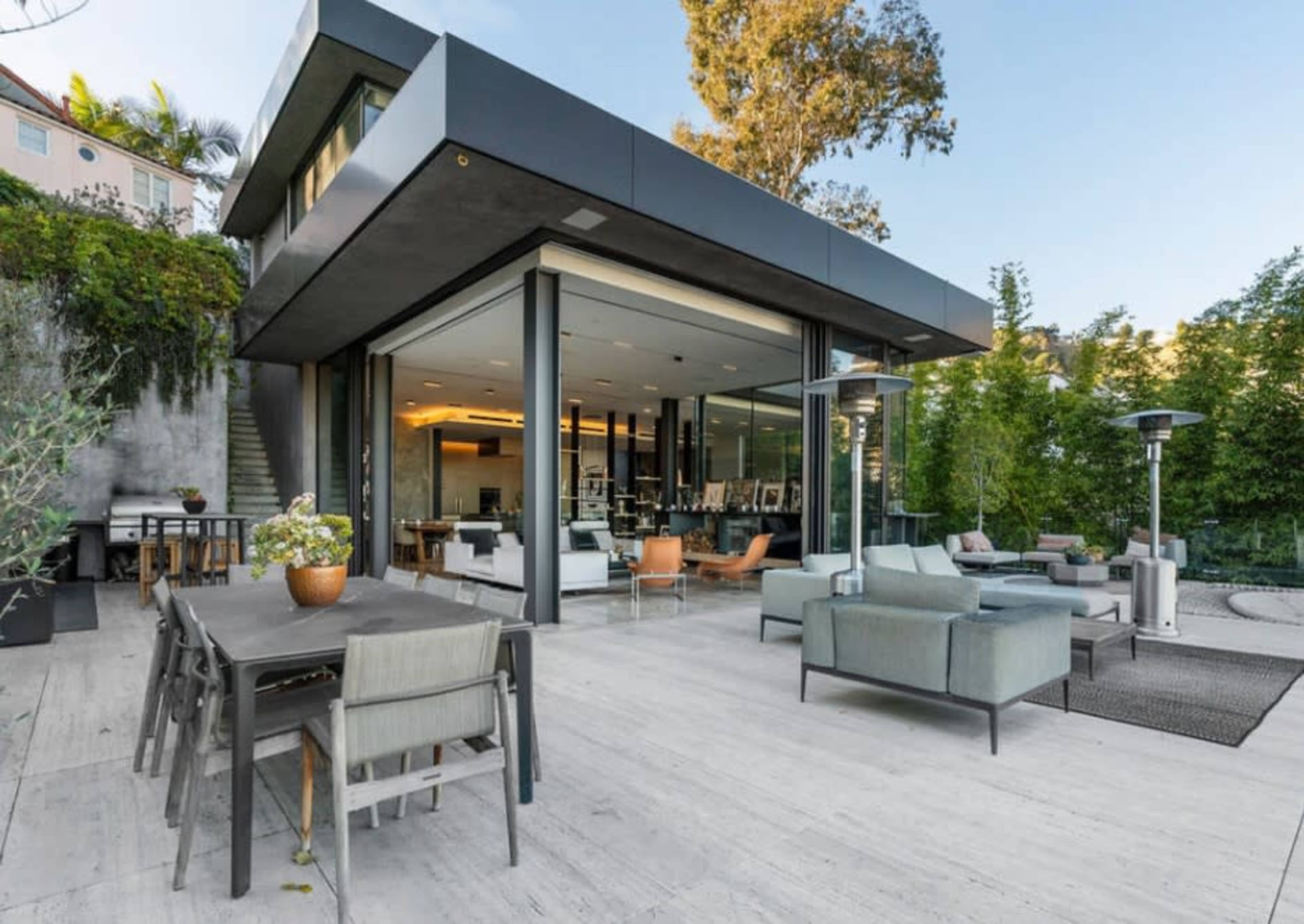 The image shows a modern house with large glass windows and a spacious outdoor terrace, featuring furniture and a dining area overlooking landscaped greenery.