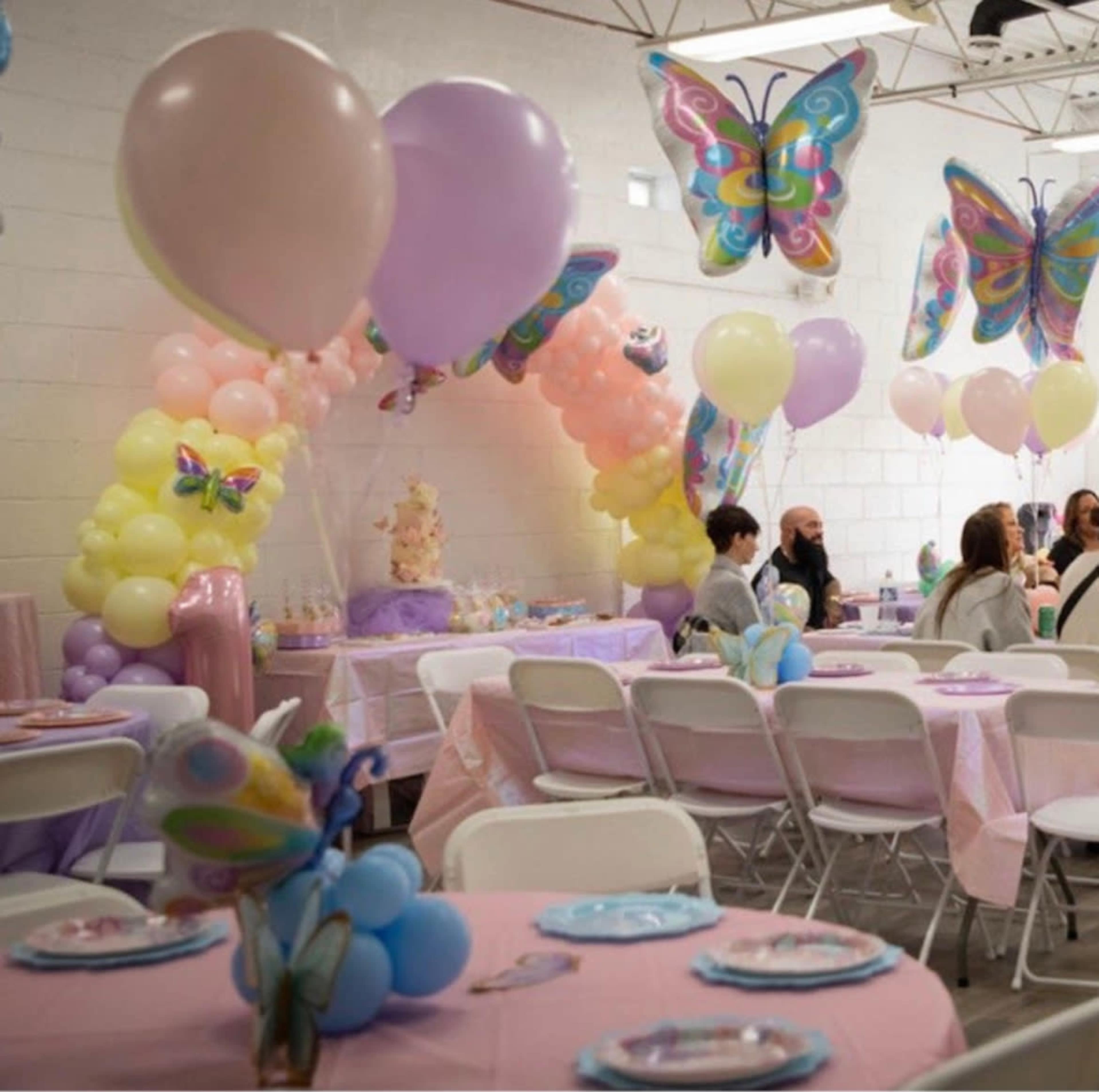 A colorful party setup features tables adorned with pastel tablecloths, butterfly decorations, and balloon arrangements in a decorated indoor space.