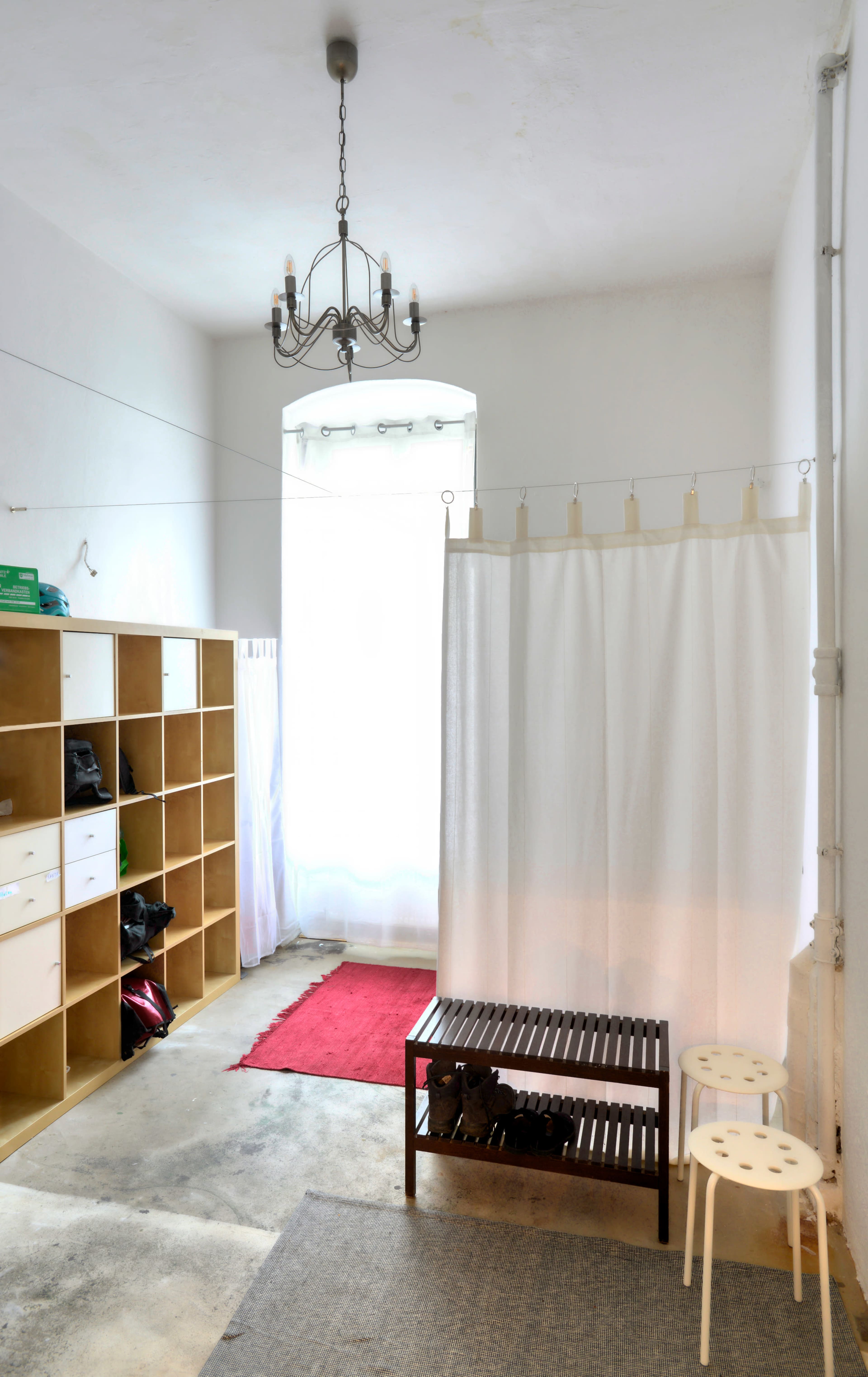 A minimalist interior space featuring a window with sheer curtains, a red rug, a wooden bench, two small white stools, and shelving units along the wall.