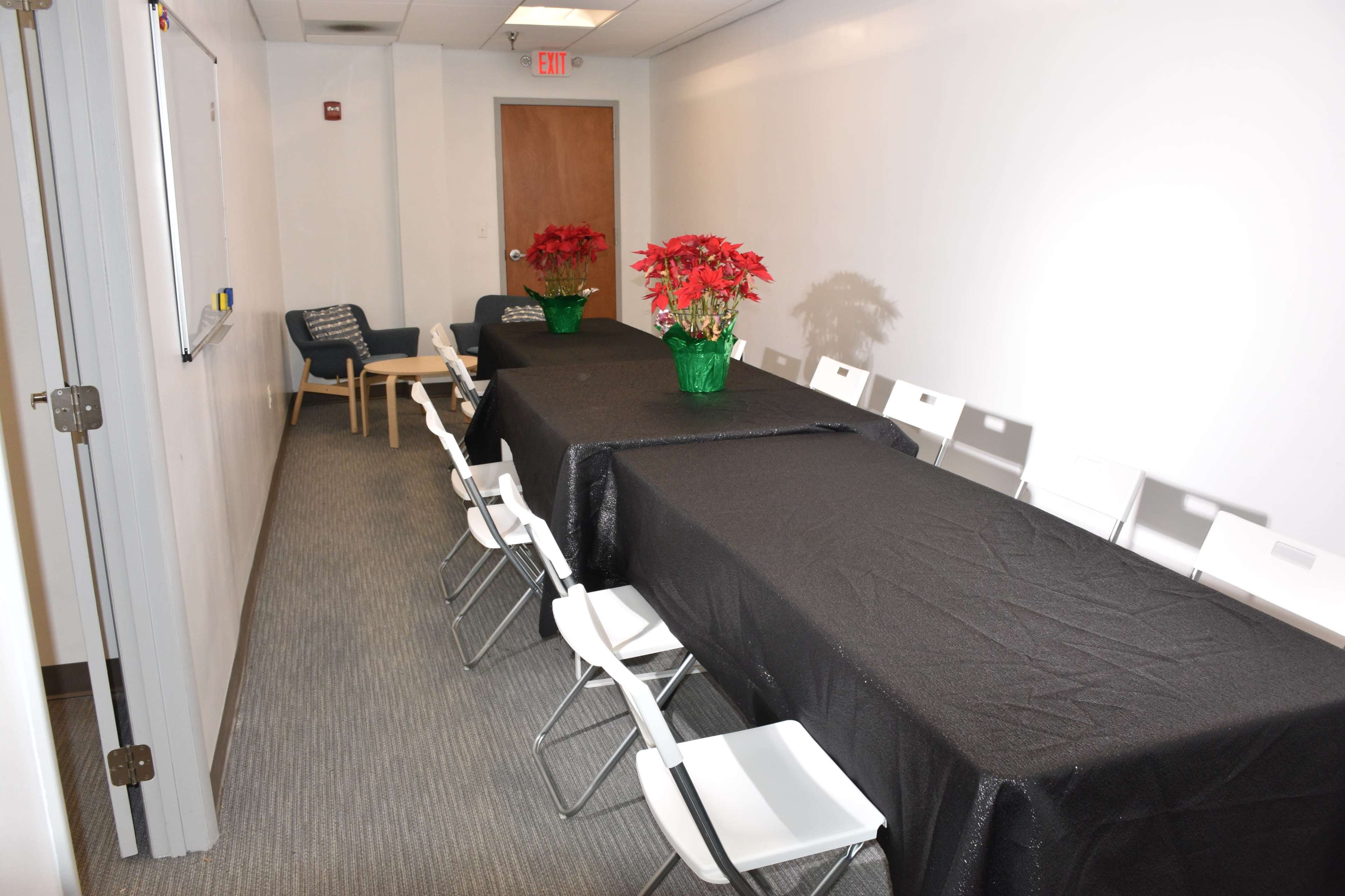 A long black-covered table with white chairs is arranged in a narrow meeting room, decorated with potted poinsettias at each end.
