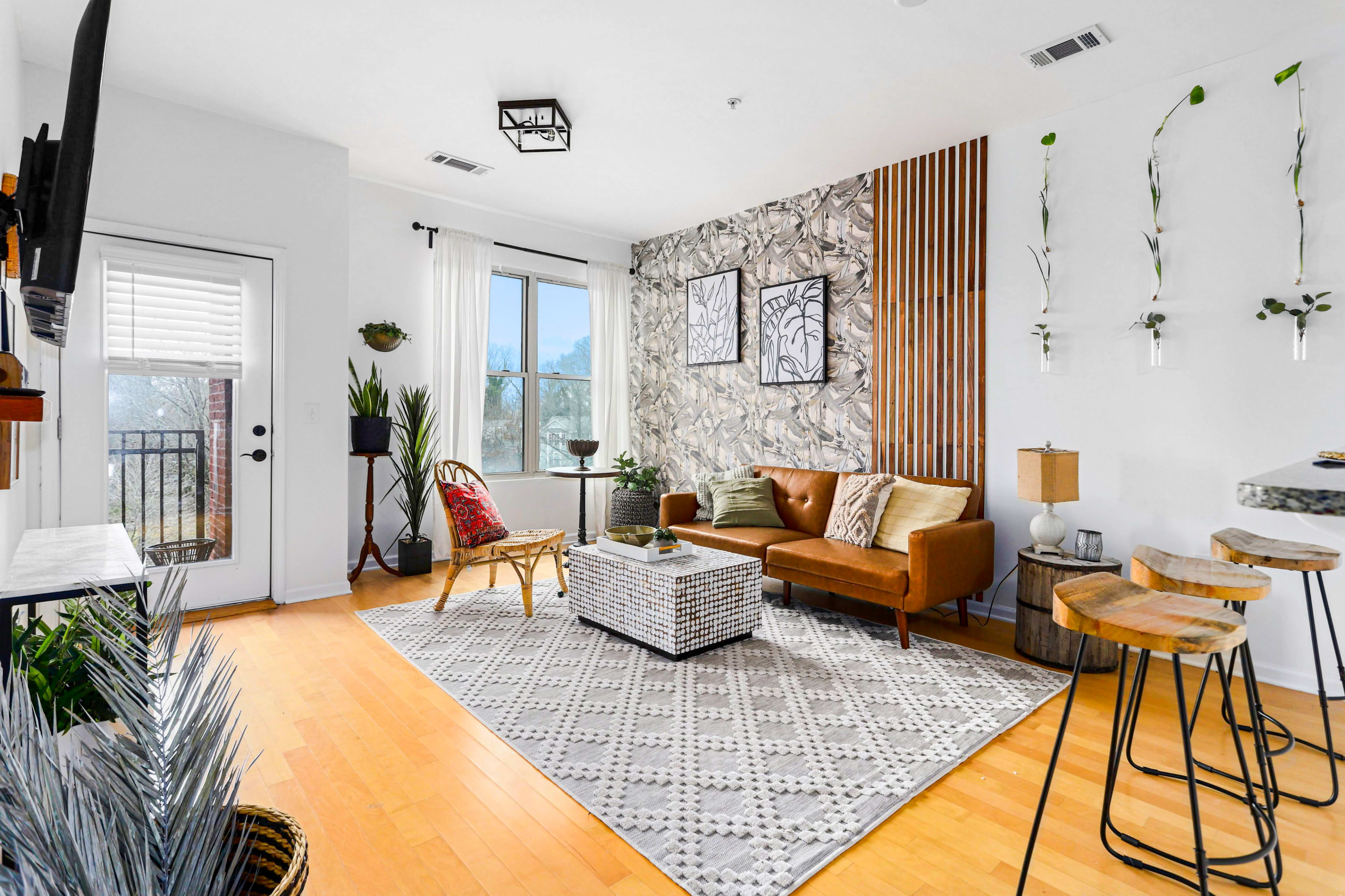 A modern living room features a brown leather couch, a decorative accent wall, and a window overlooking a balcony.