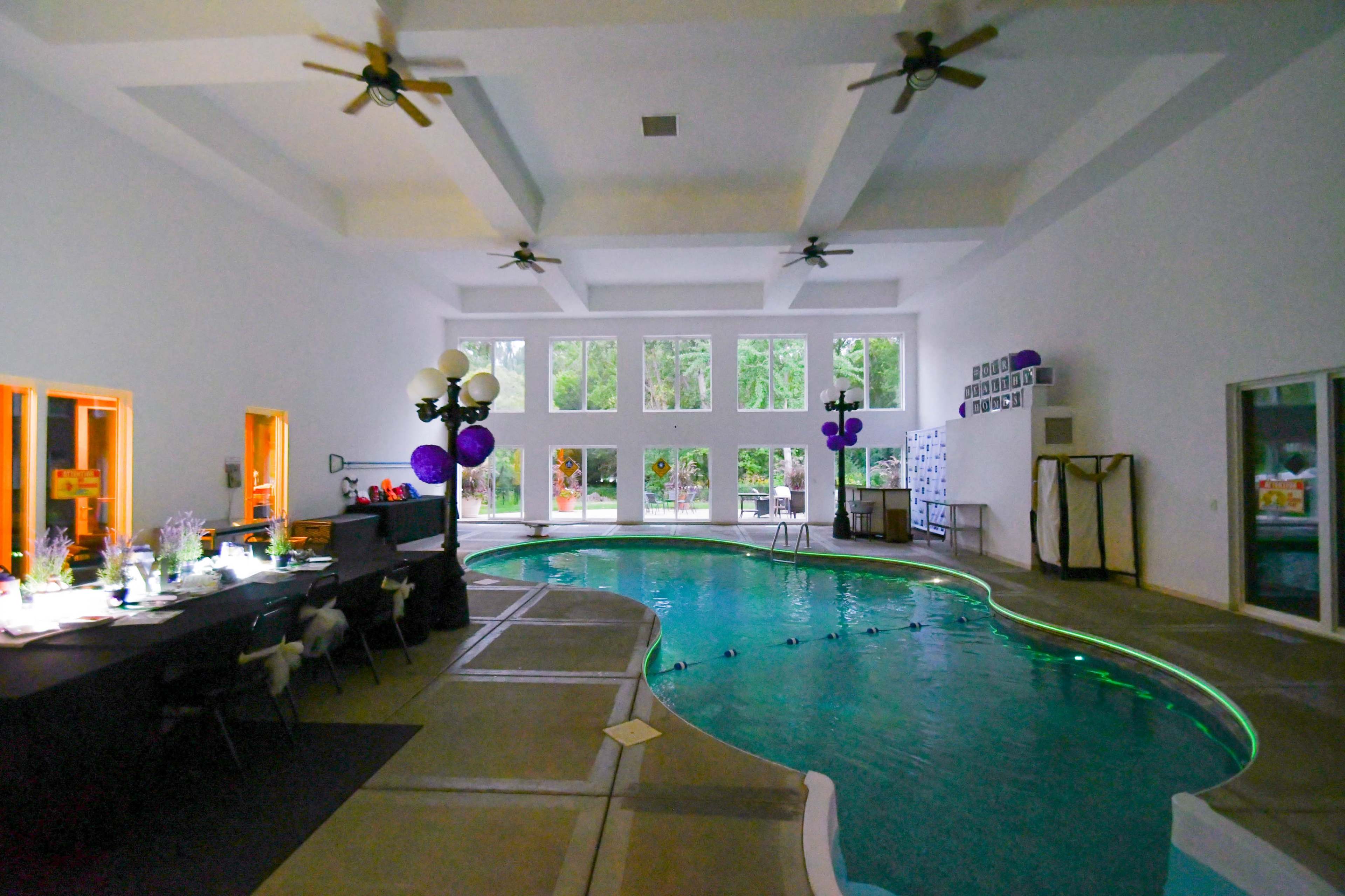 An indoor pool area with large windows, a dark table setup, and decorations in shades of purple.