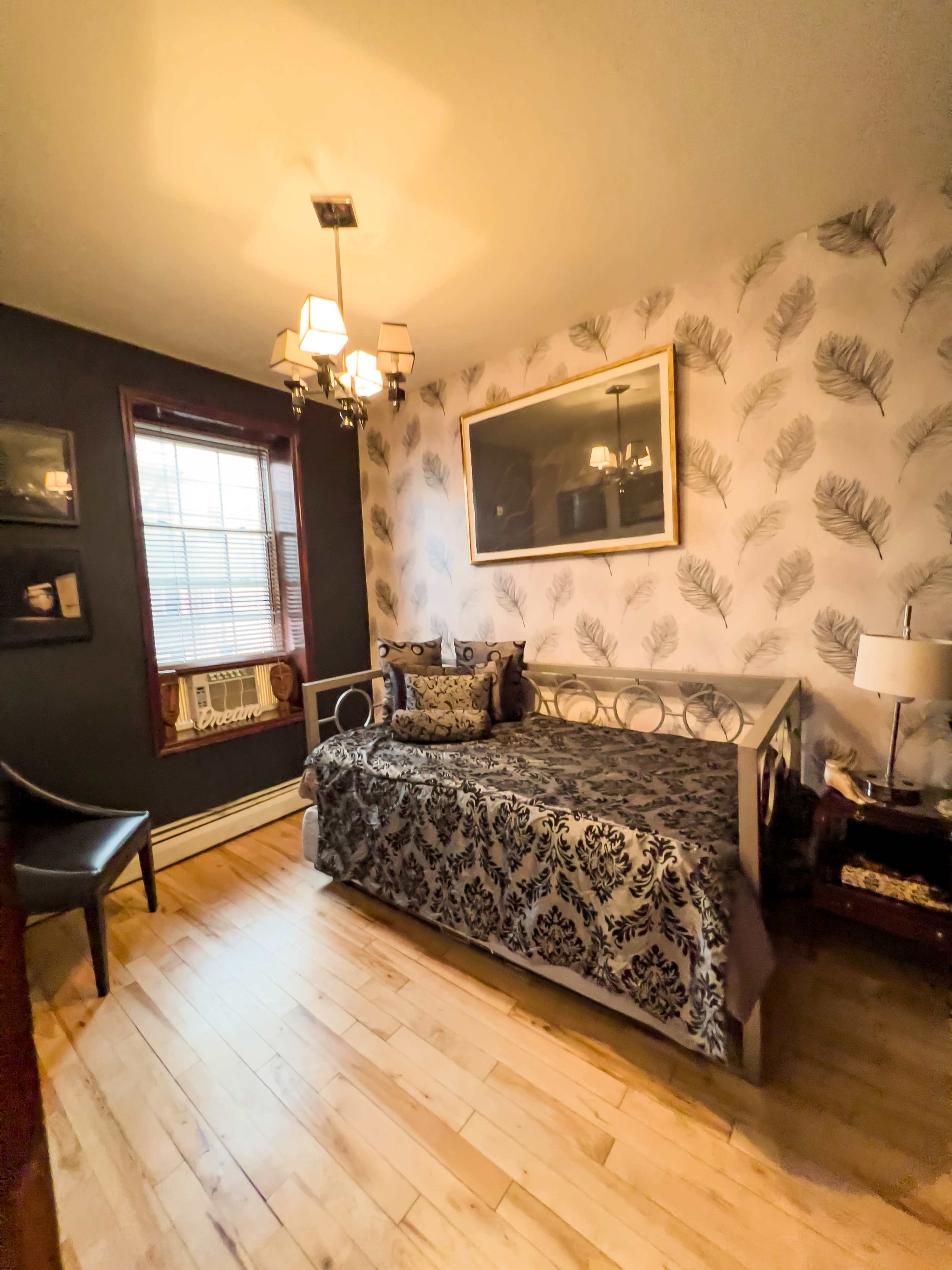 A small bedroom with a daybed, decorative pillows, and a patterned wallpaper, featuring a window and an air conditioner unit.