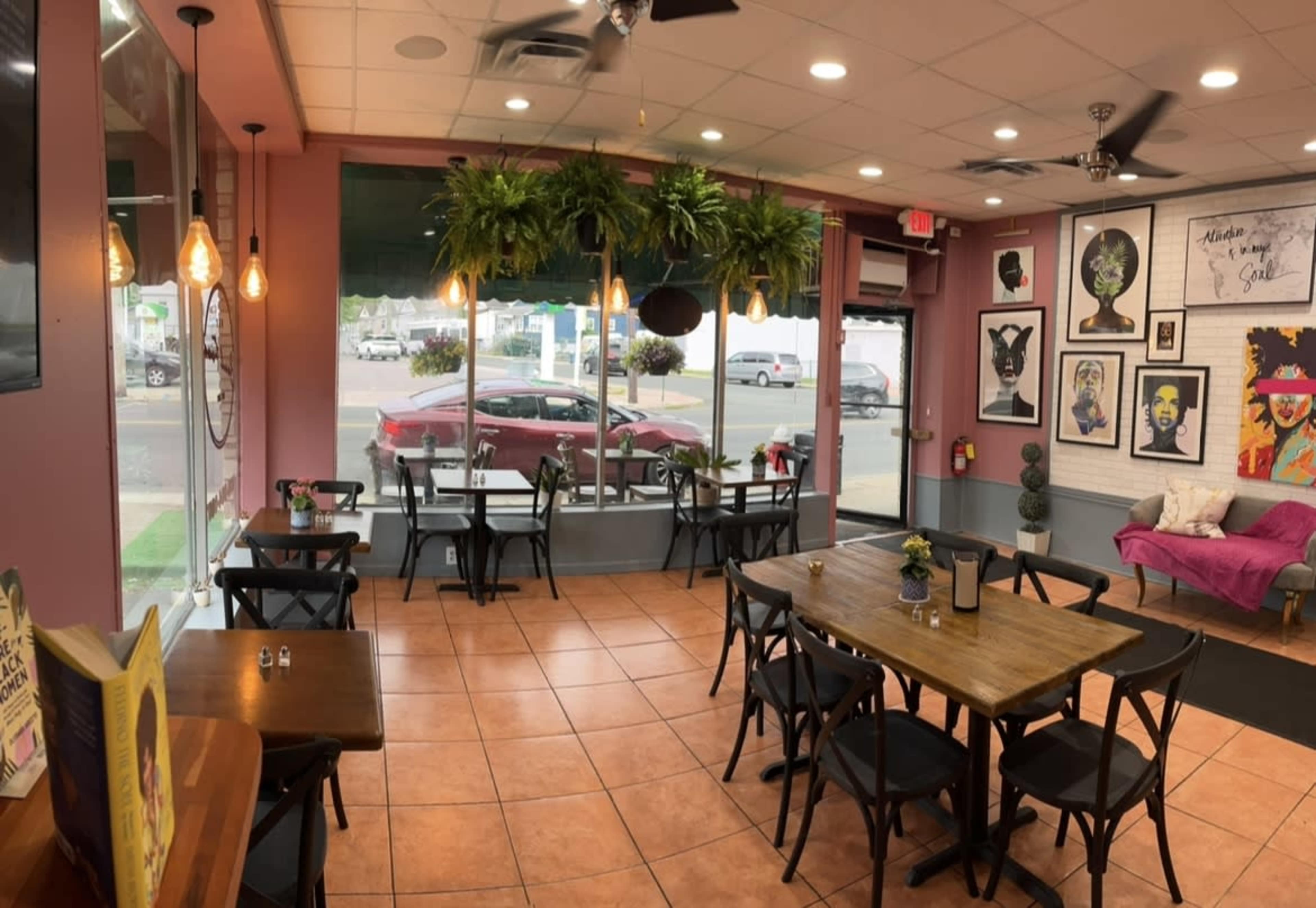 A bright cafe features wooden tables, potted plants hanging from the ceiling, and colorful wall art near large windows.