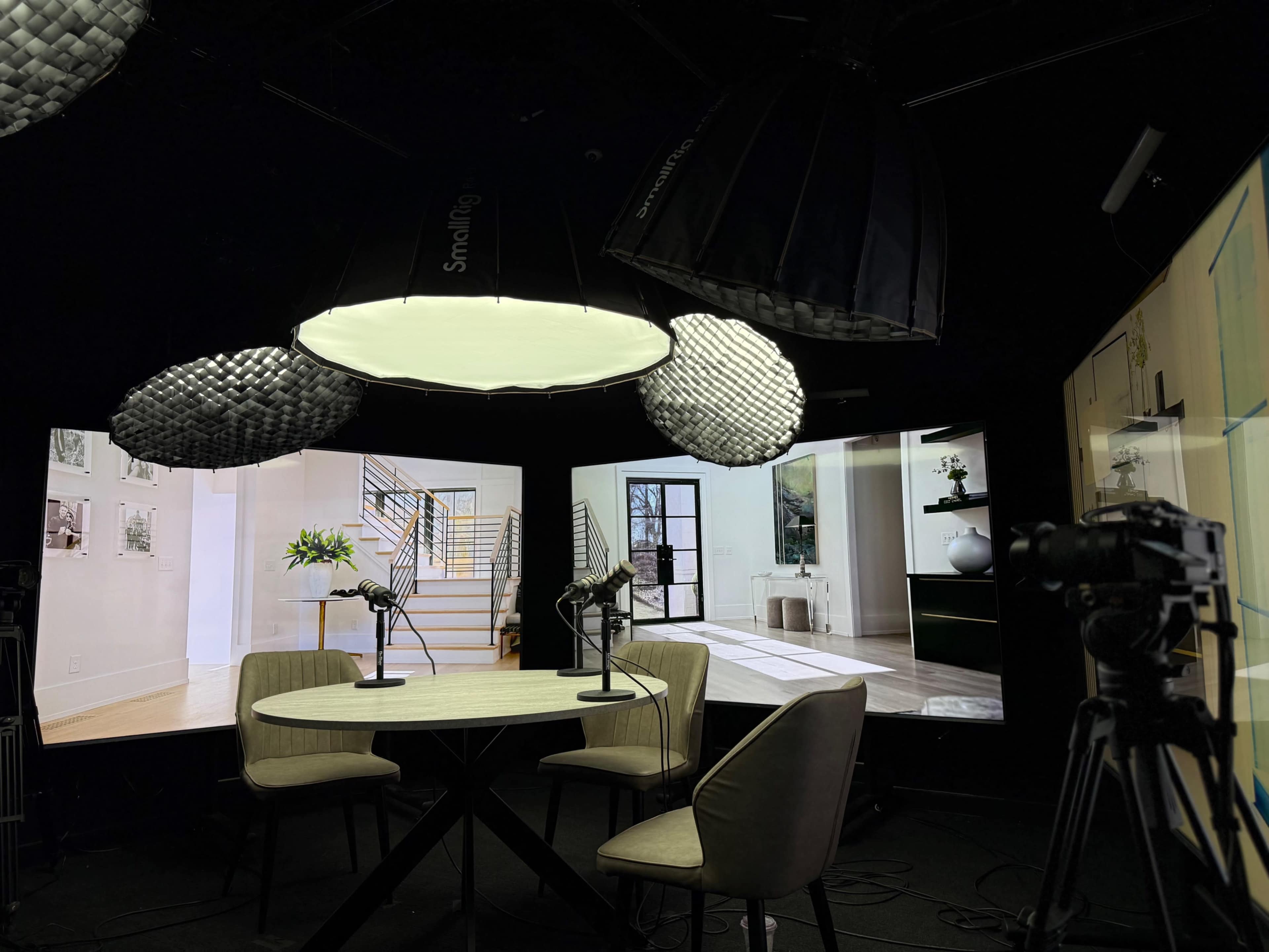 A modern studio setup features a round table with two chairs, surrounded by large screens displaying interior designs and illuminated by unique lighting fixtures.