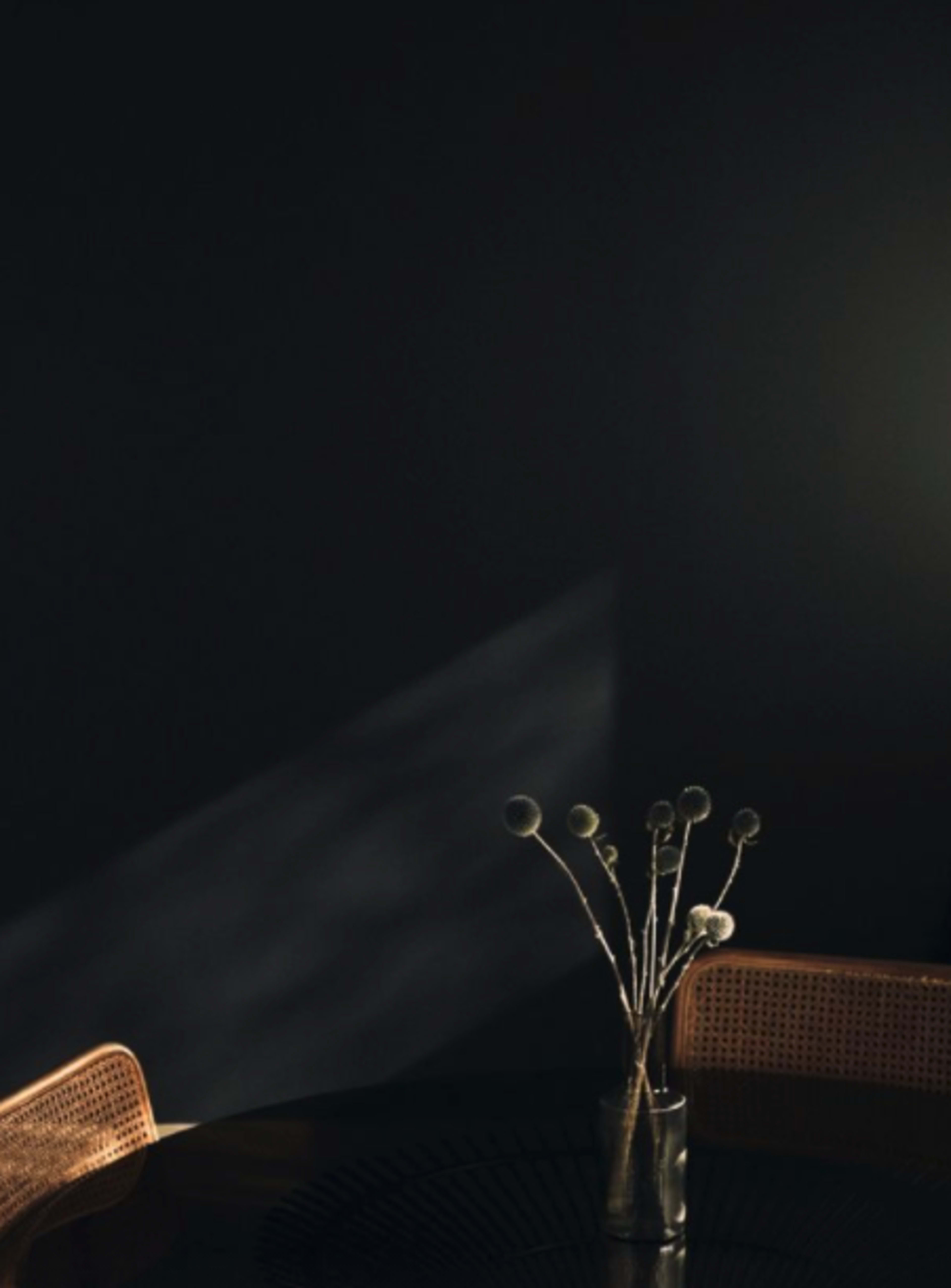 A vase with dried flowers is placed on a round table under dim lighting, against a dark wall.