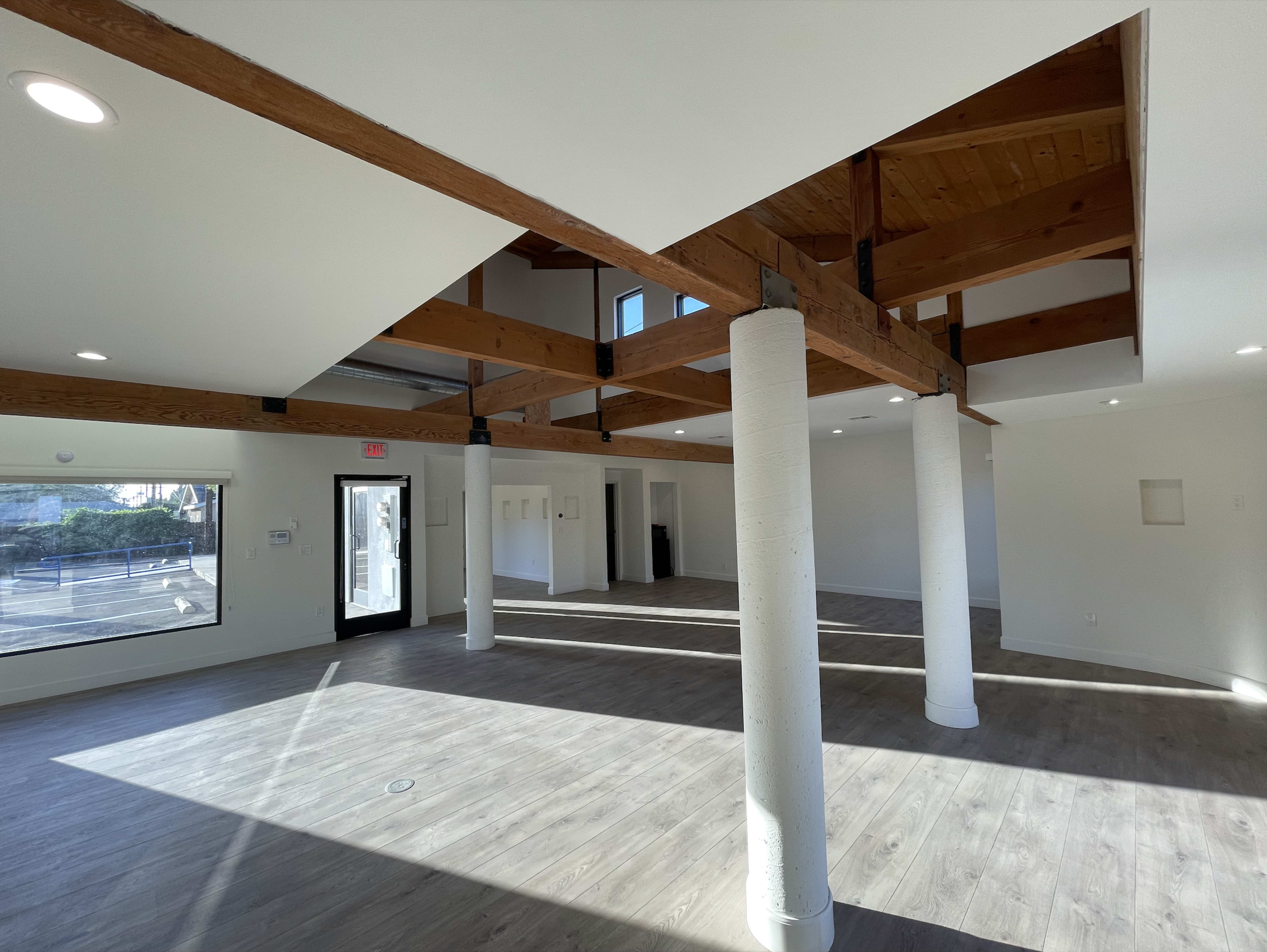 The image shows a spacious interior room with wooden beams, tall ceilings, and large windows, featuring a polished floor and columns.