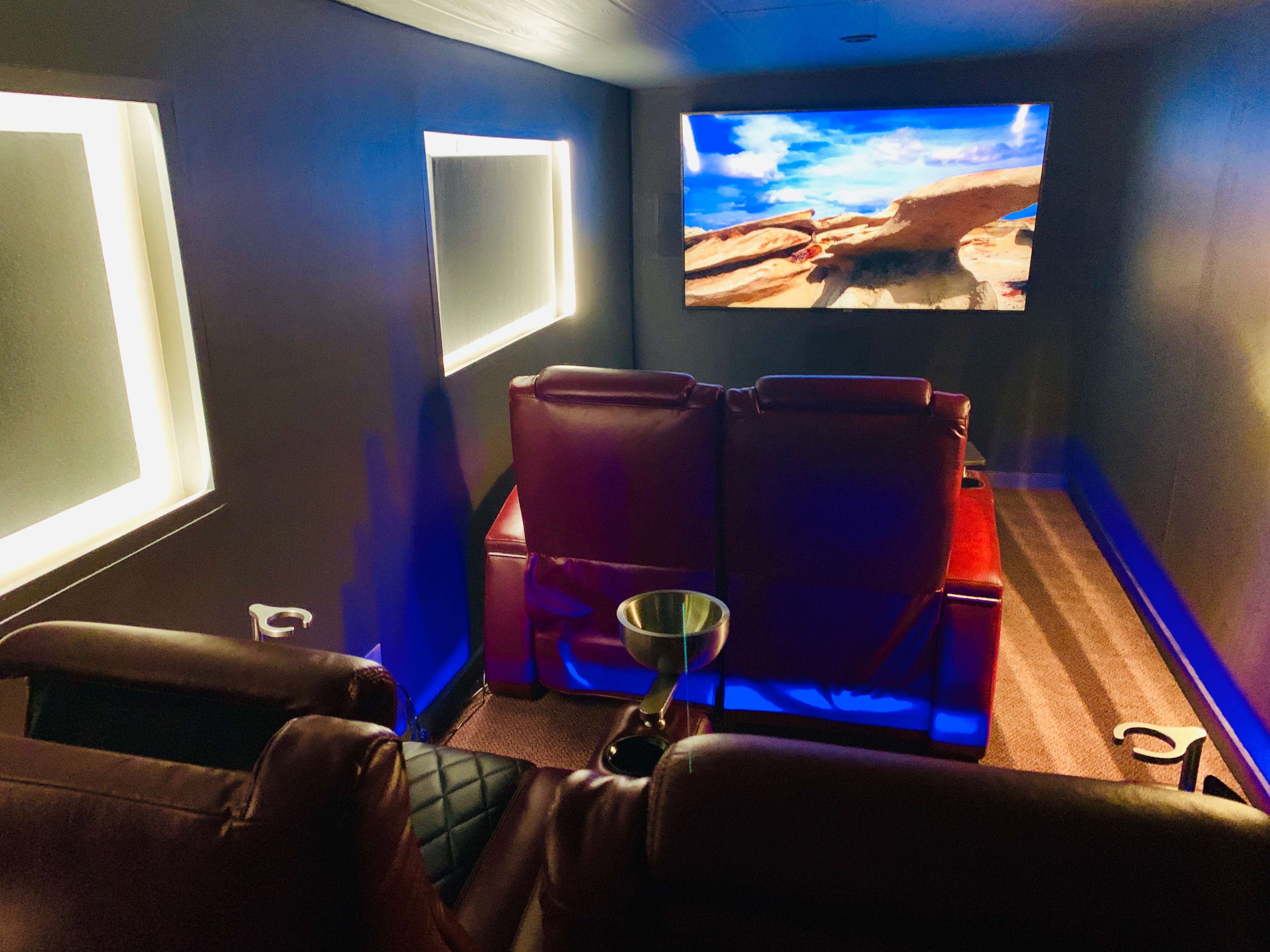 A dimly lit home theater features two red recliner chairs facing a large screen showing a scenic landscape.