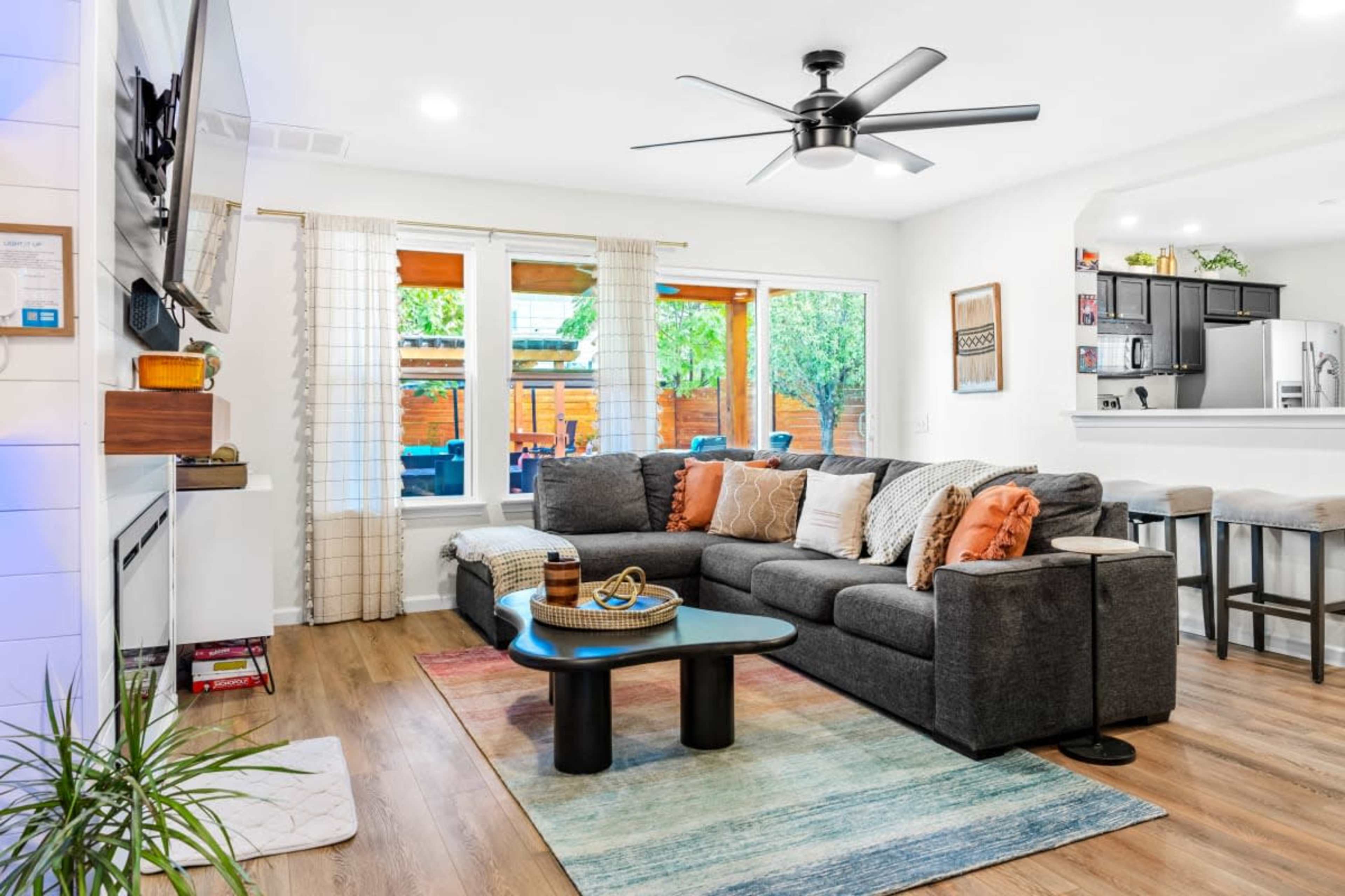A modern living room featuring a grey sectional sofa with colorful throw pillows, a coffee table, a fan, and large windows that open to an outdoor patio.