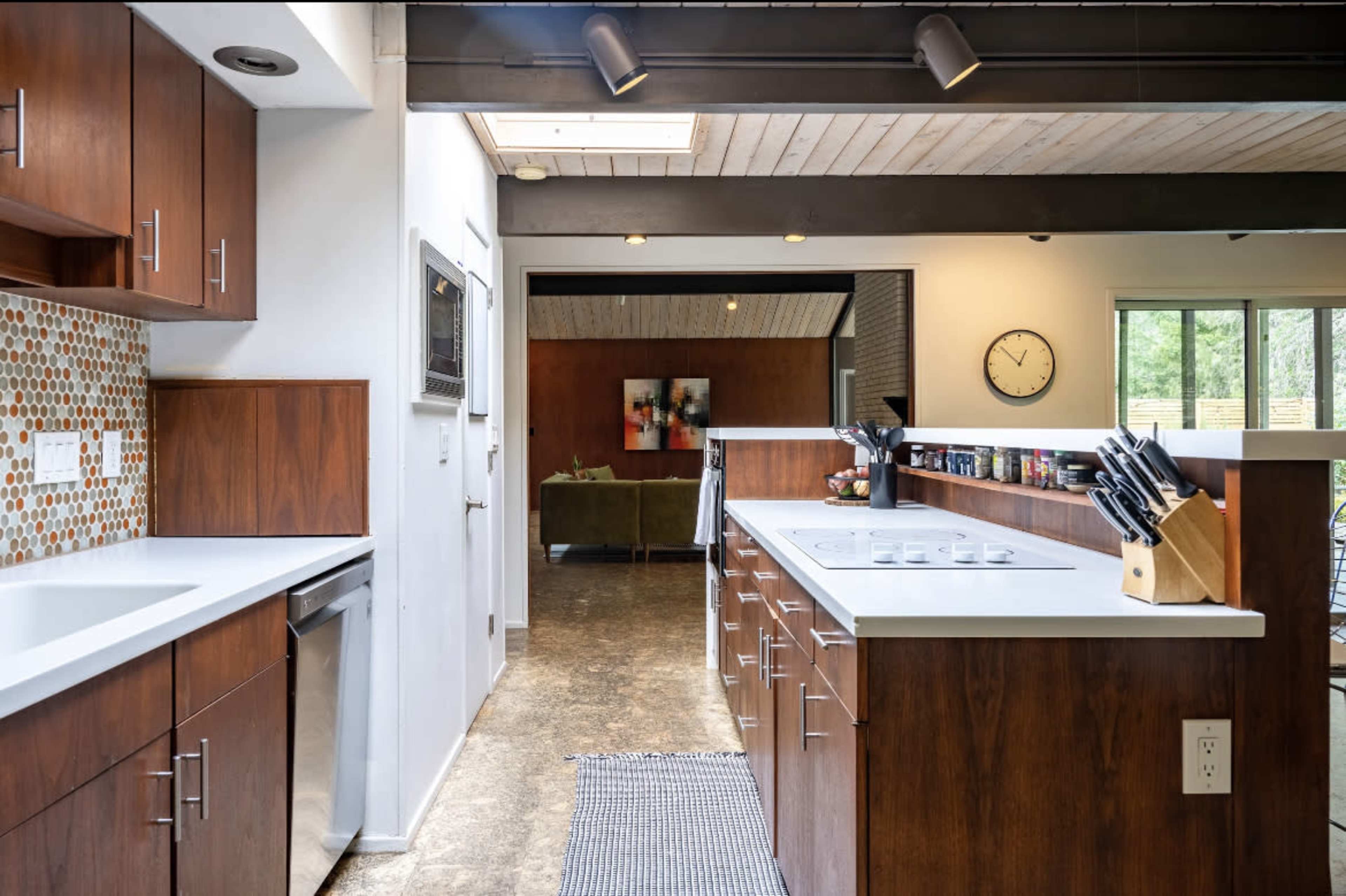 A modern kitchen features wooden cabinetry, a white countertop, and a mosaic tile backsplash, with an open view to a living area.
