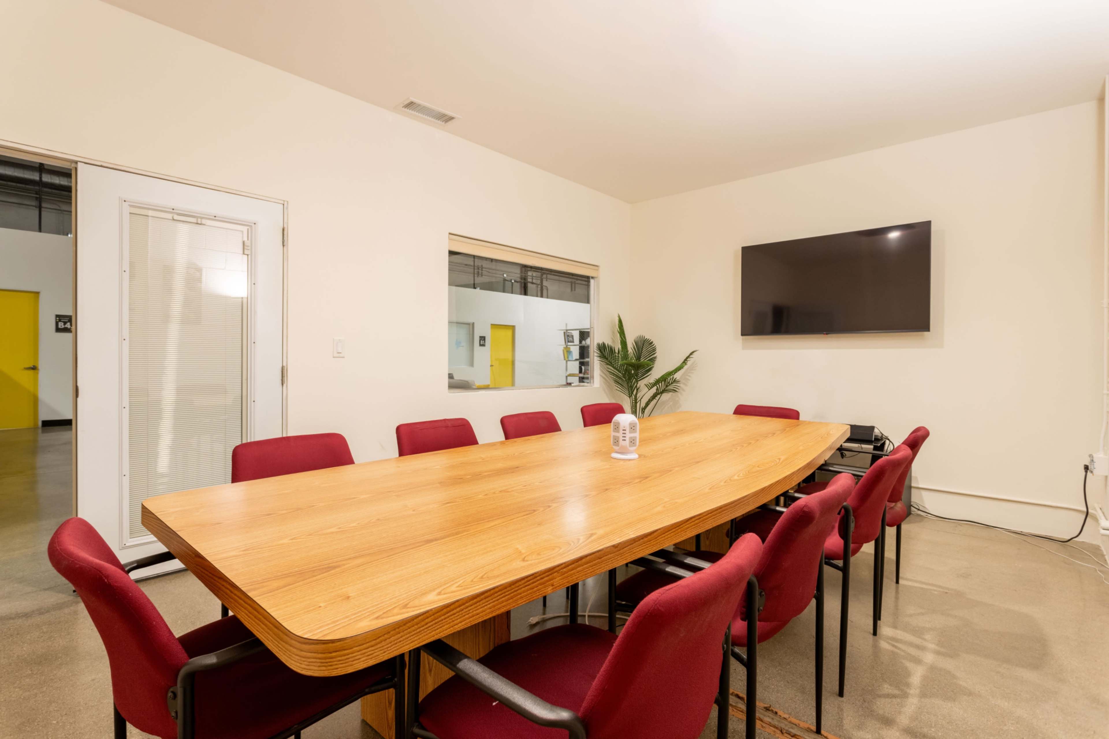 The image shows a conference room with a long wooden table surrounded by red chairs, a television on the wall, and a potted plant in the corner.