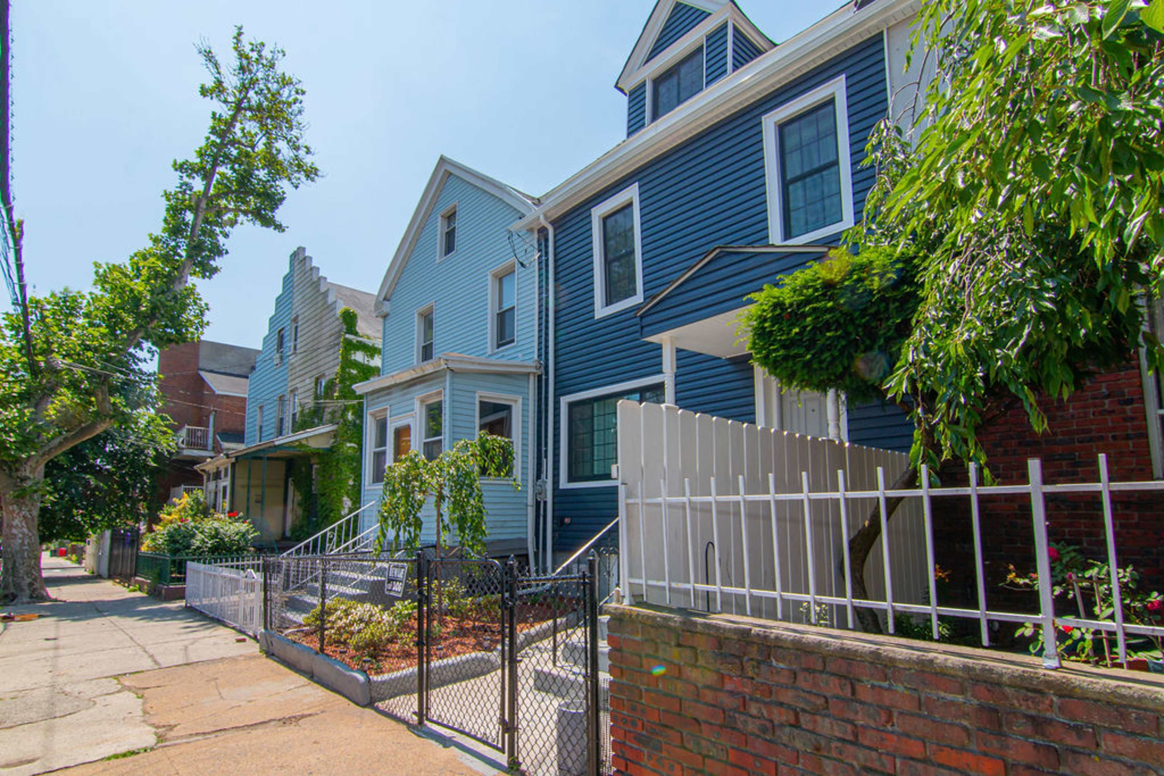 The image shows a row of residential houses, featuring blue and gray exteriors, with a white picket fence and green landscaping in the front yards.