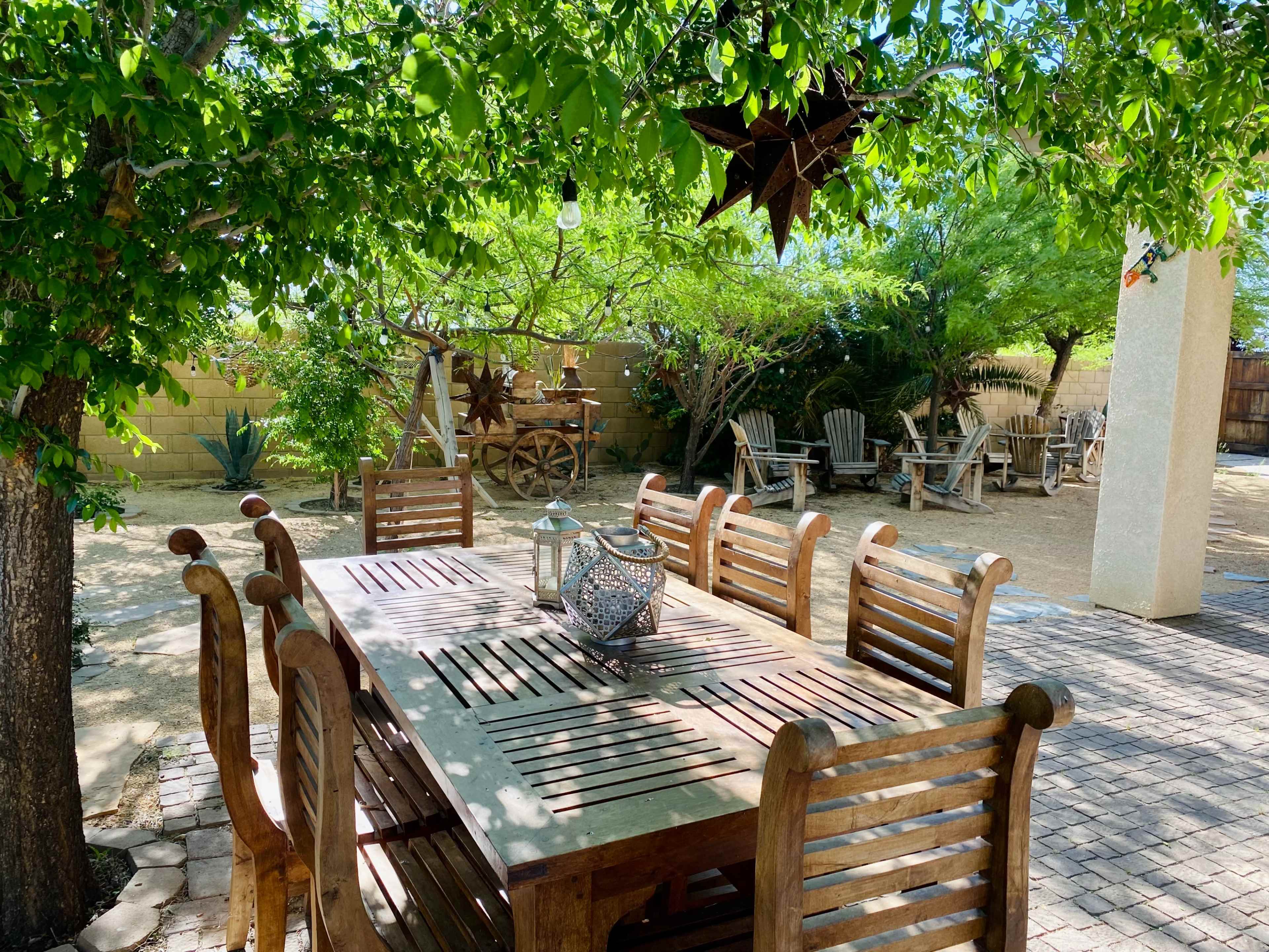 A wooden dining table surrounded by eight chairs is set beneath a leafy tree in a spacious outdoor area with additional seating in the background.