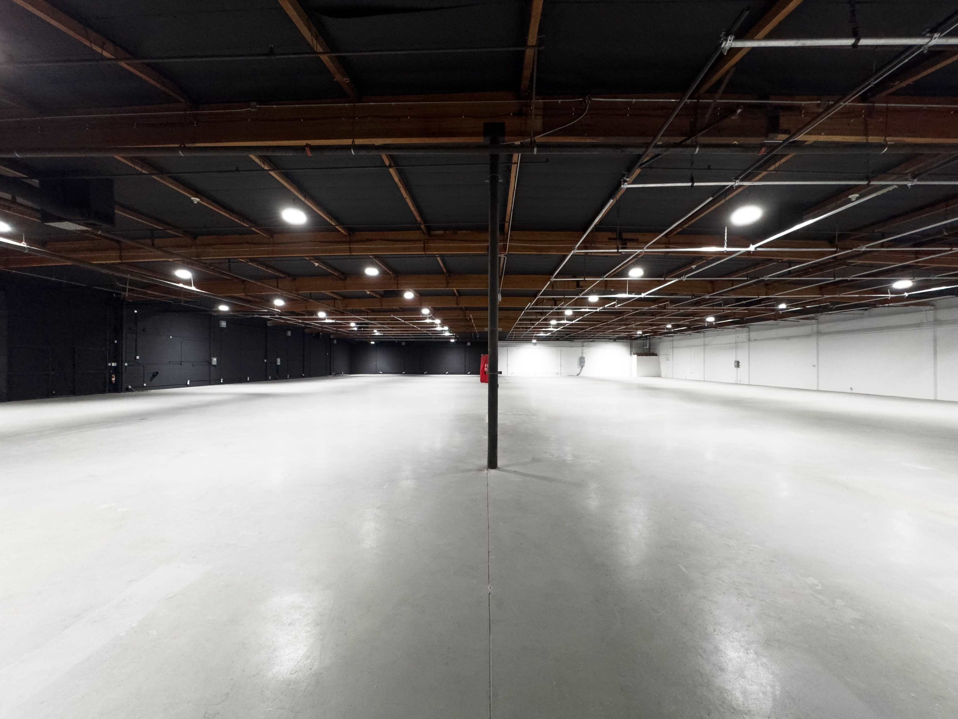 A spacious industrial warehouse features concrete flooring and exposed wooden beams, with bright overhead lighting illuminating the empty interior.