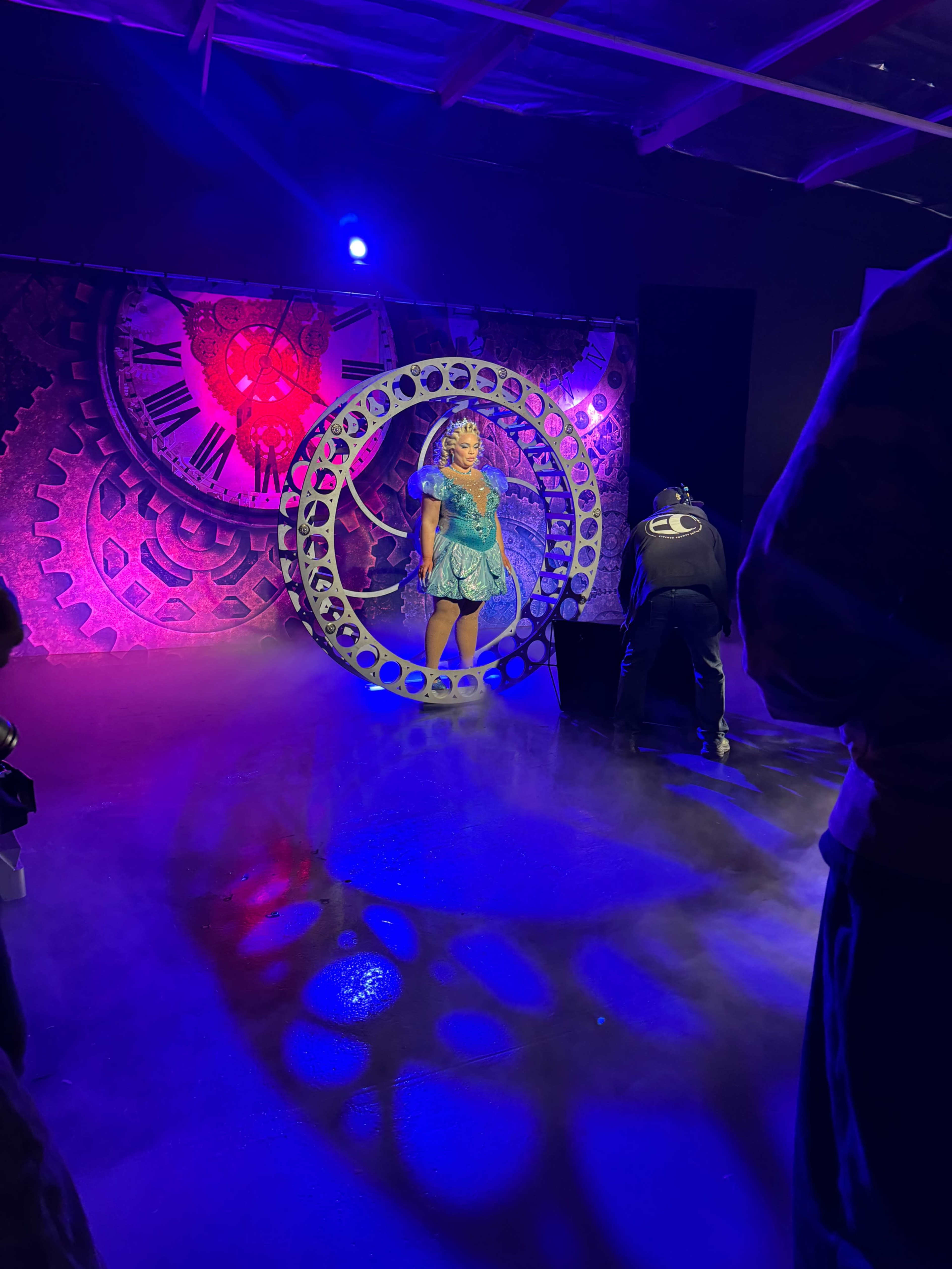 A performer in a blue costume stands inside a large circular frame on a brightly lit stage, with a backdrop featuring gears and a clock.