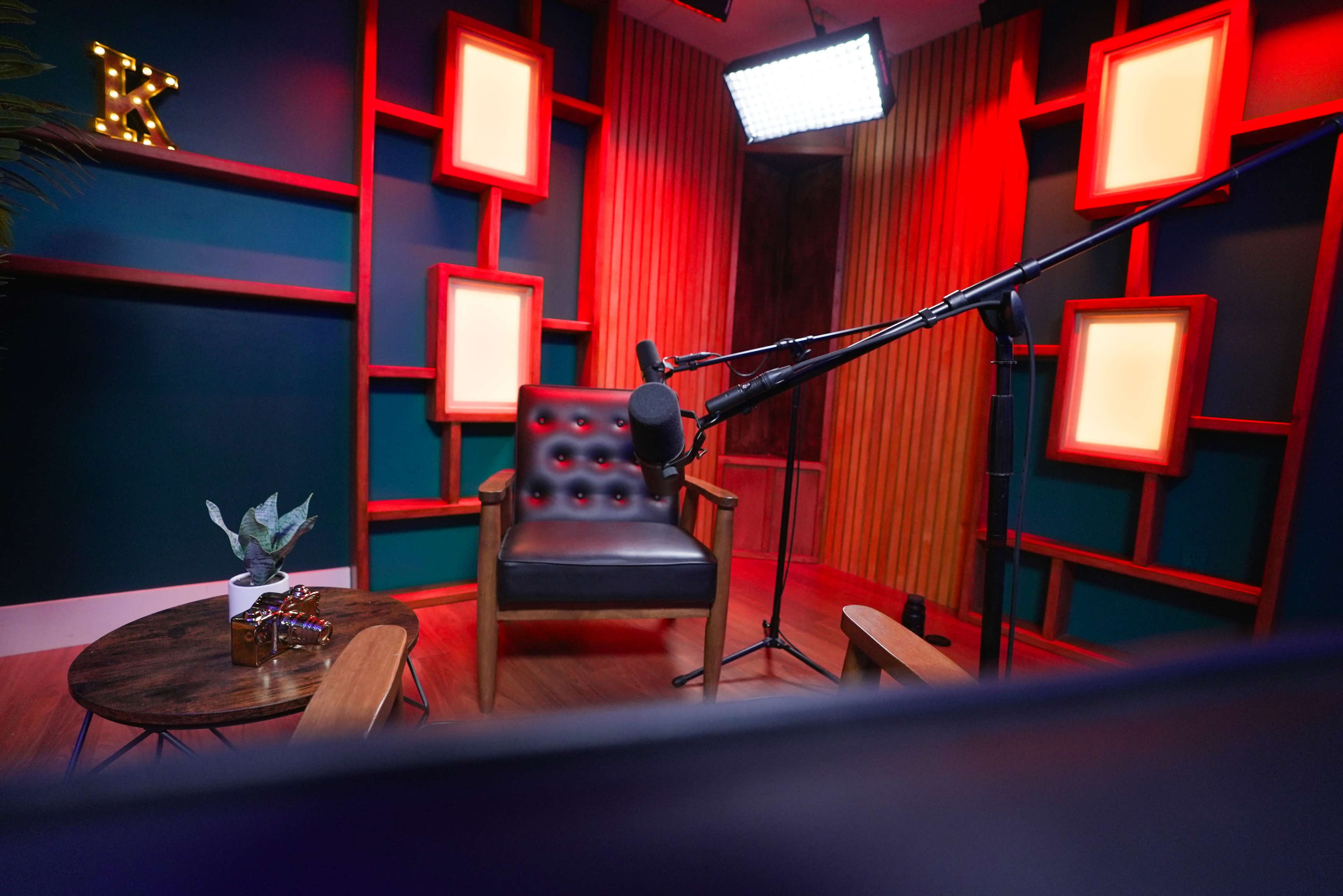 The image shows a neatly arranged podcast or interview setup featuring a black chair, a microphone on a stand, and a coffee table with a small plant and decorative items, all set against a backdrop of illuminated frames and colorful walls.