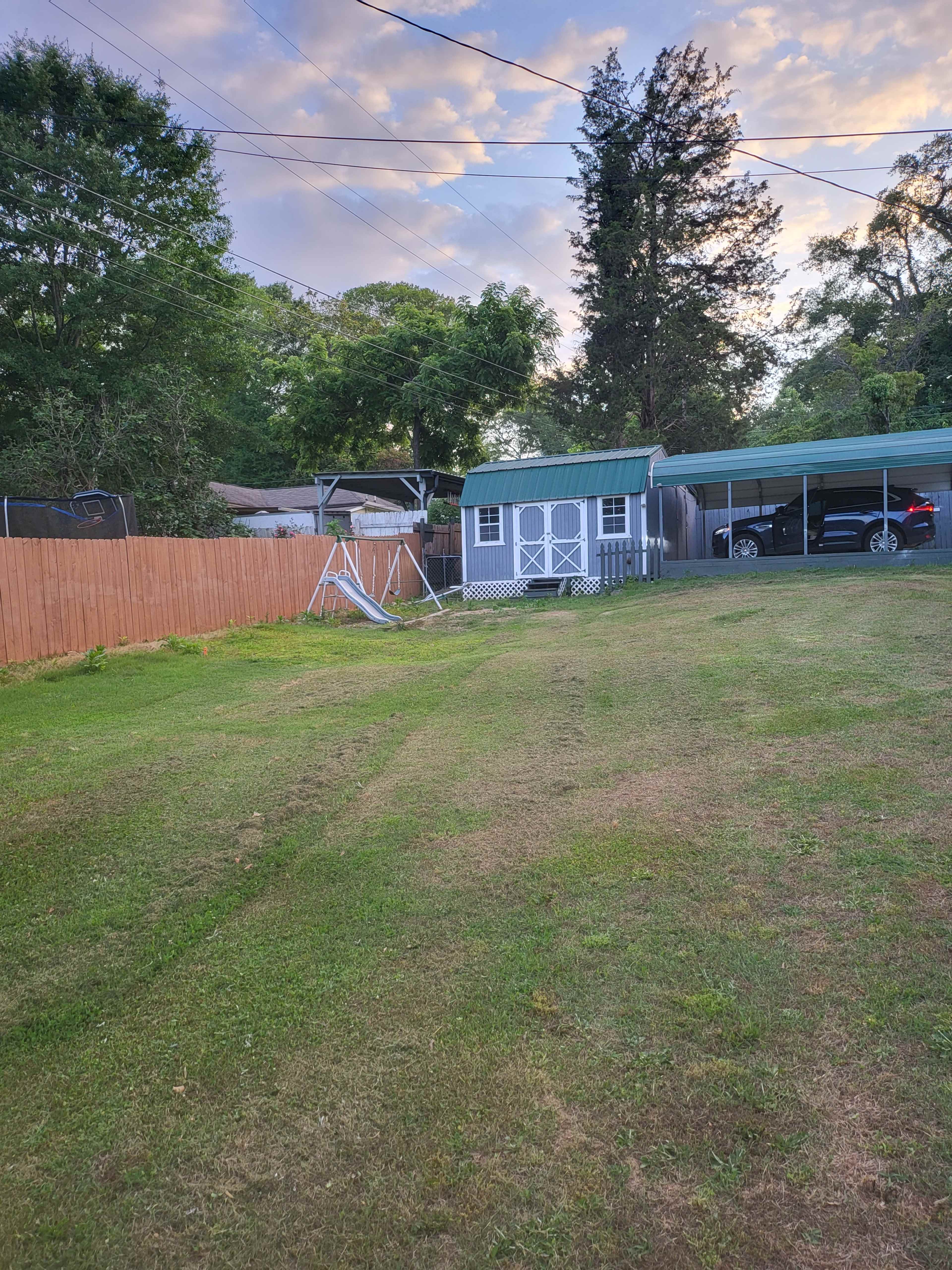 A grassy yard with a storage shed, a carport, and a swing set against a backdrop of trees and a cloudy sky.