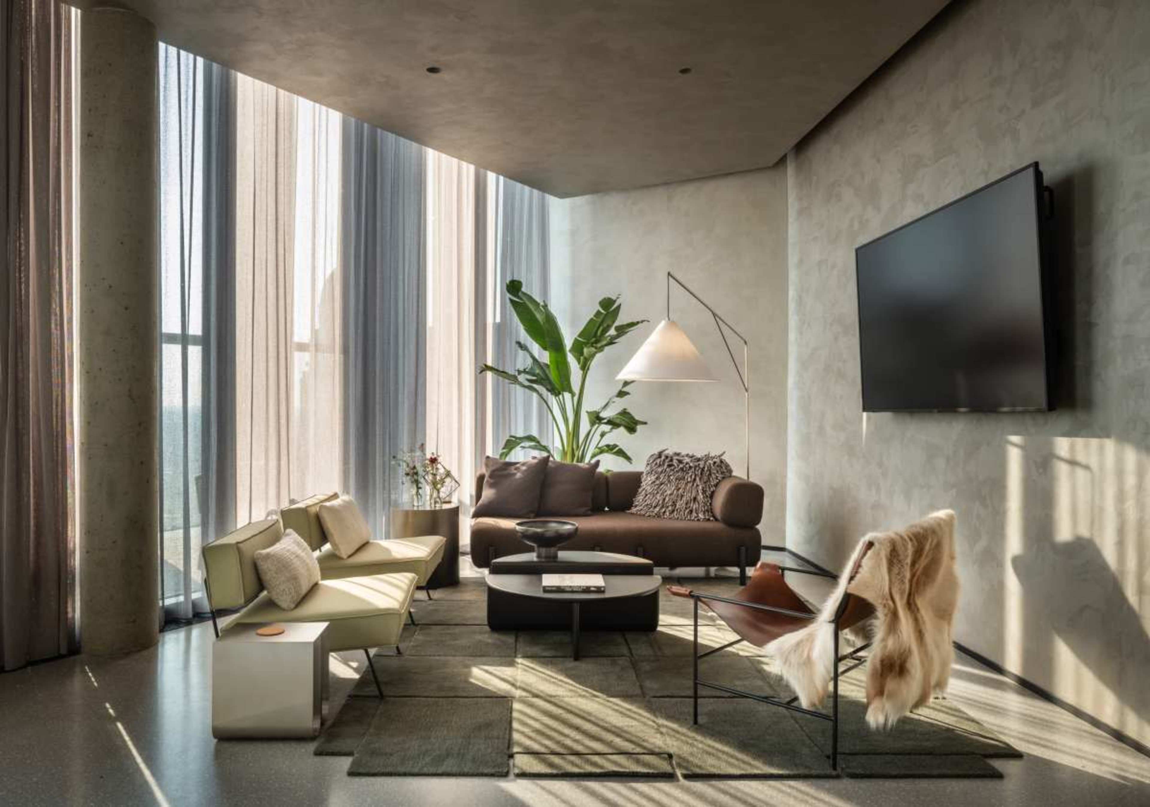 The image shows a modern living room with a brown sofa, a white armchair, a round coffee table, and large windows draped with sheer curtains, allowing natural light to fill the space.
