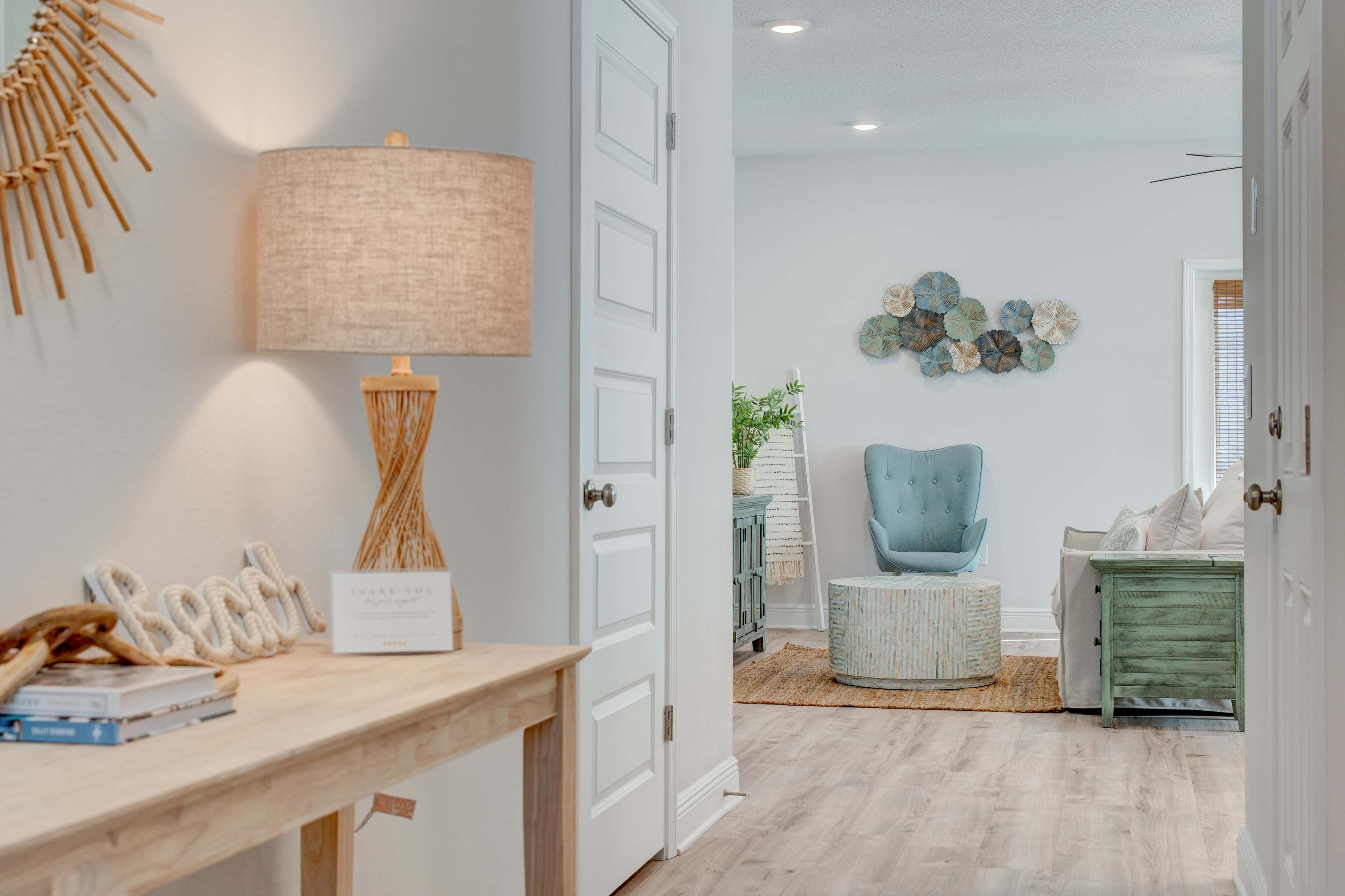 The scene features a well-lit hallway leading to a cozy living area furnished with a blue accent chair, a green cabinet, and decorative wall art.