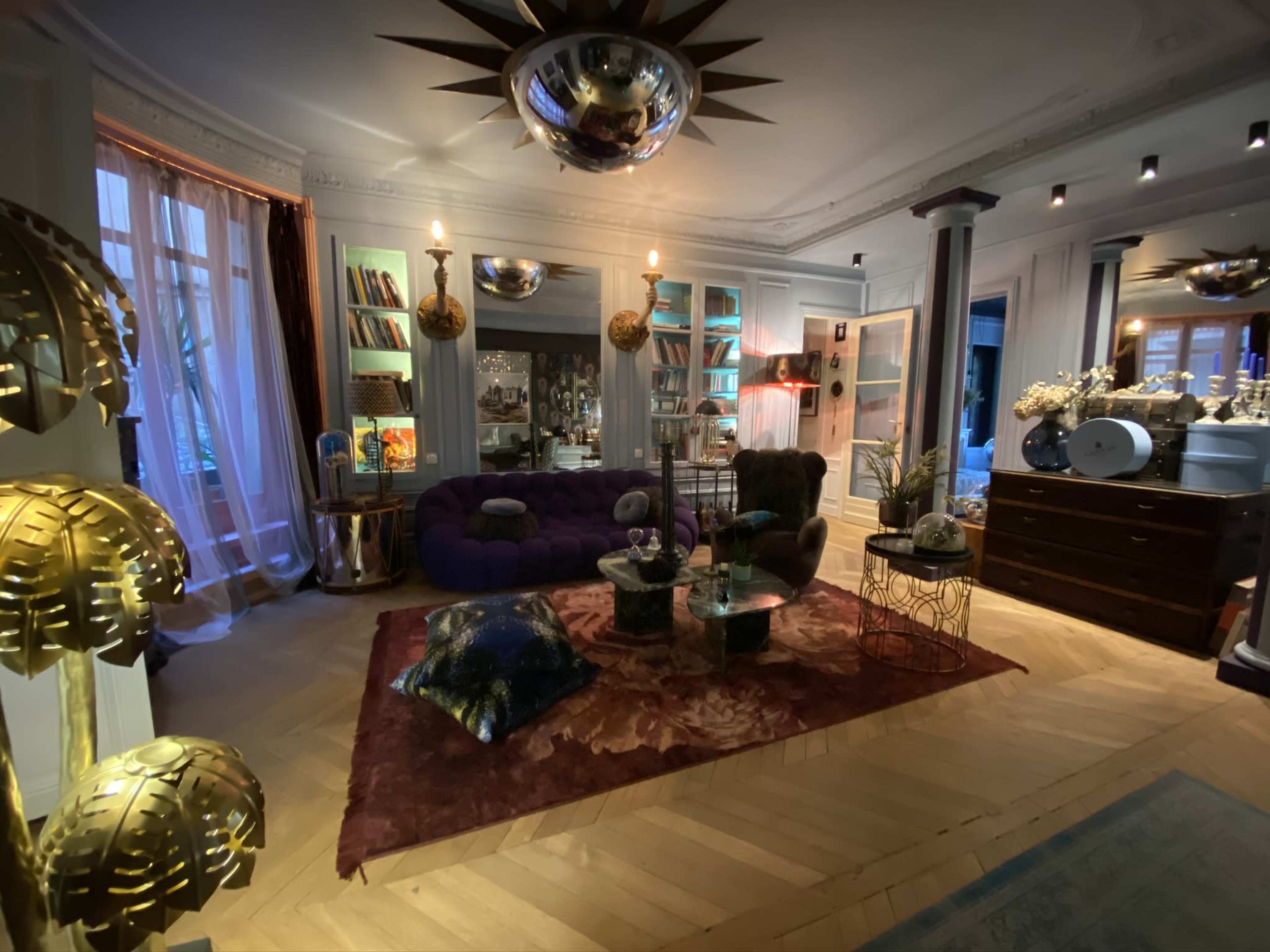 The scene features a stylish living room with a purple sofa, elegant lighting fixtures, a coffee table, and various decorative elements on a wooden floor.