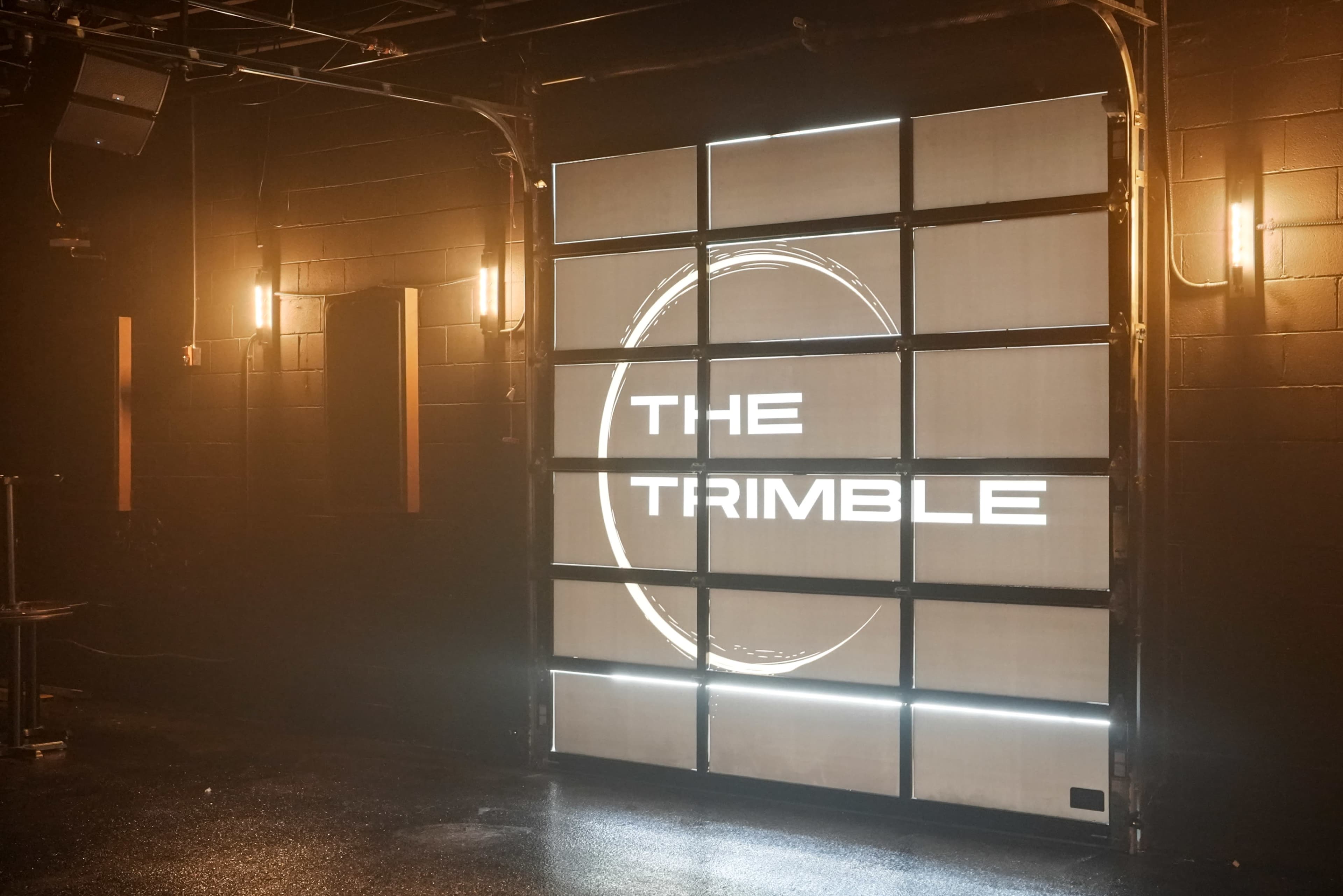 The image shows a dimly lit venue with a large illuminated sign reading "THE TRIMBLE" on a panel made up of rectangular sections.