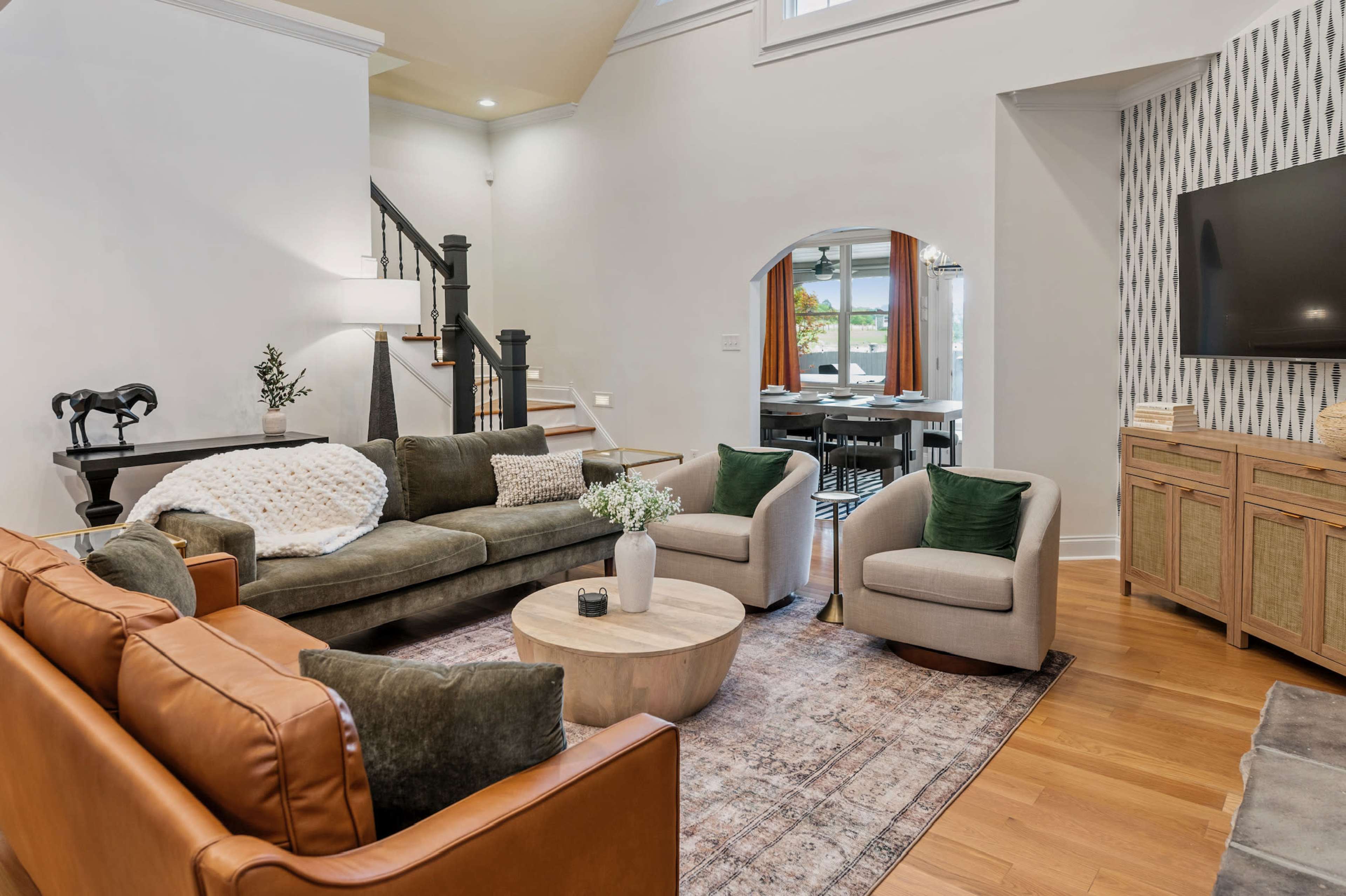 The image shows a modern living room with a mix of seating options, including a brown leather sofa and light-colored armchairs, arranged around a round coffee table on a patterned rug.