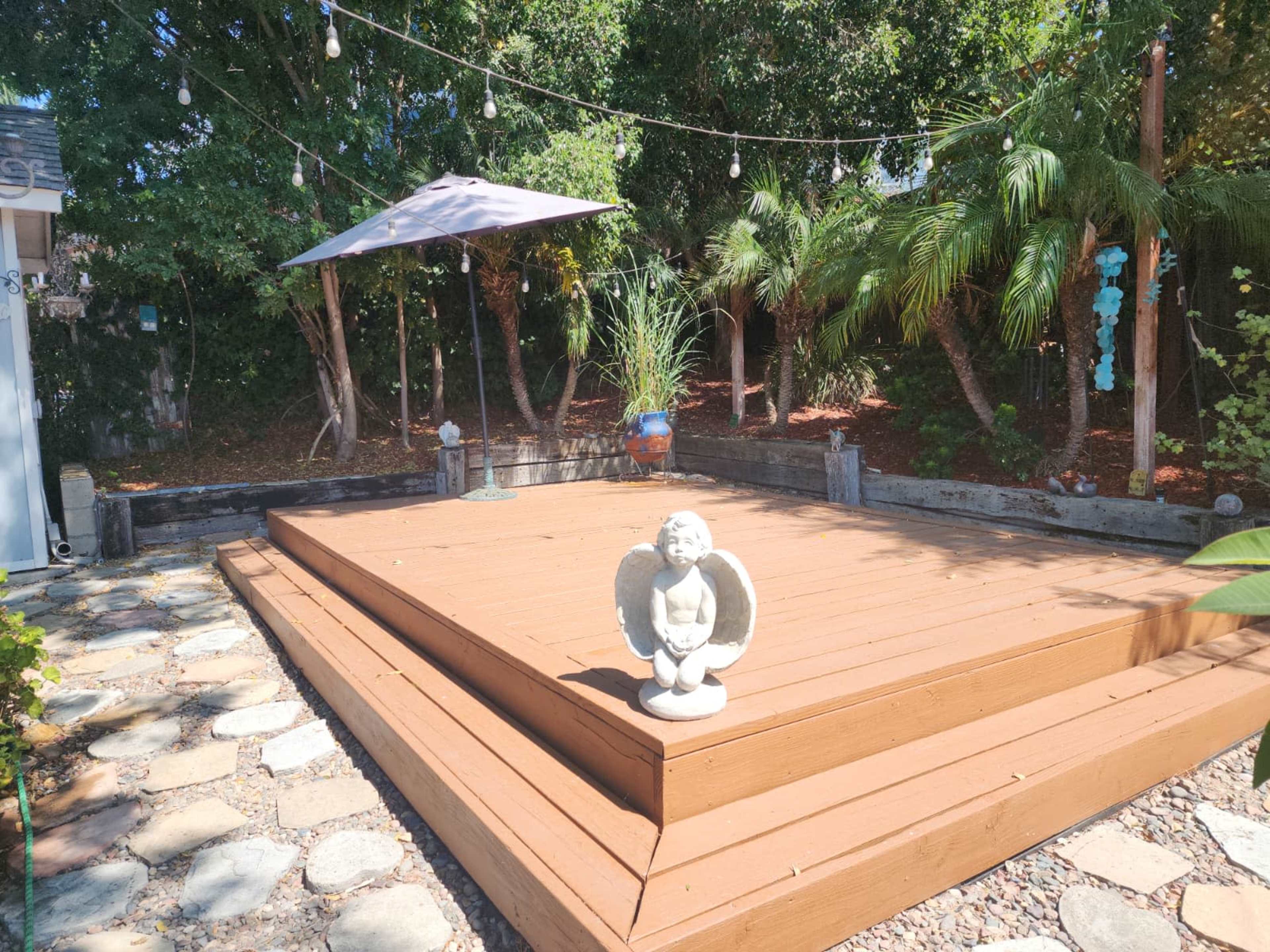 The image shows a wooden deck in a backyard surrounded by greenery, featuring a small angel statue and a canopy umbrella above.
