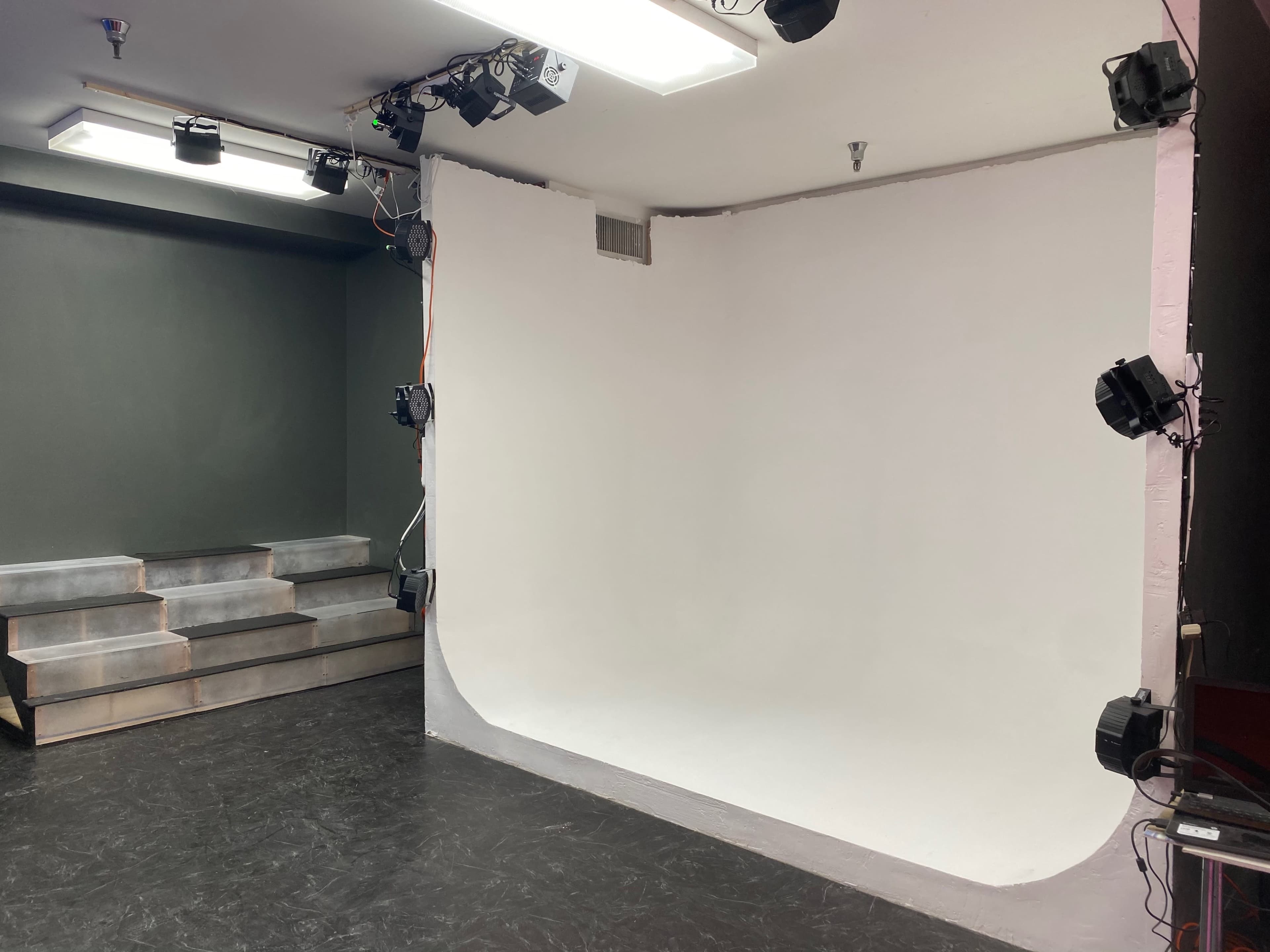 A photography studio with a curved white backdrop, multiple lighting sets mounted on the walls, and tiered steps in the background.