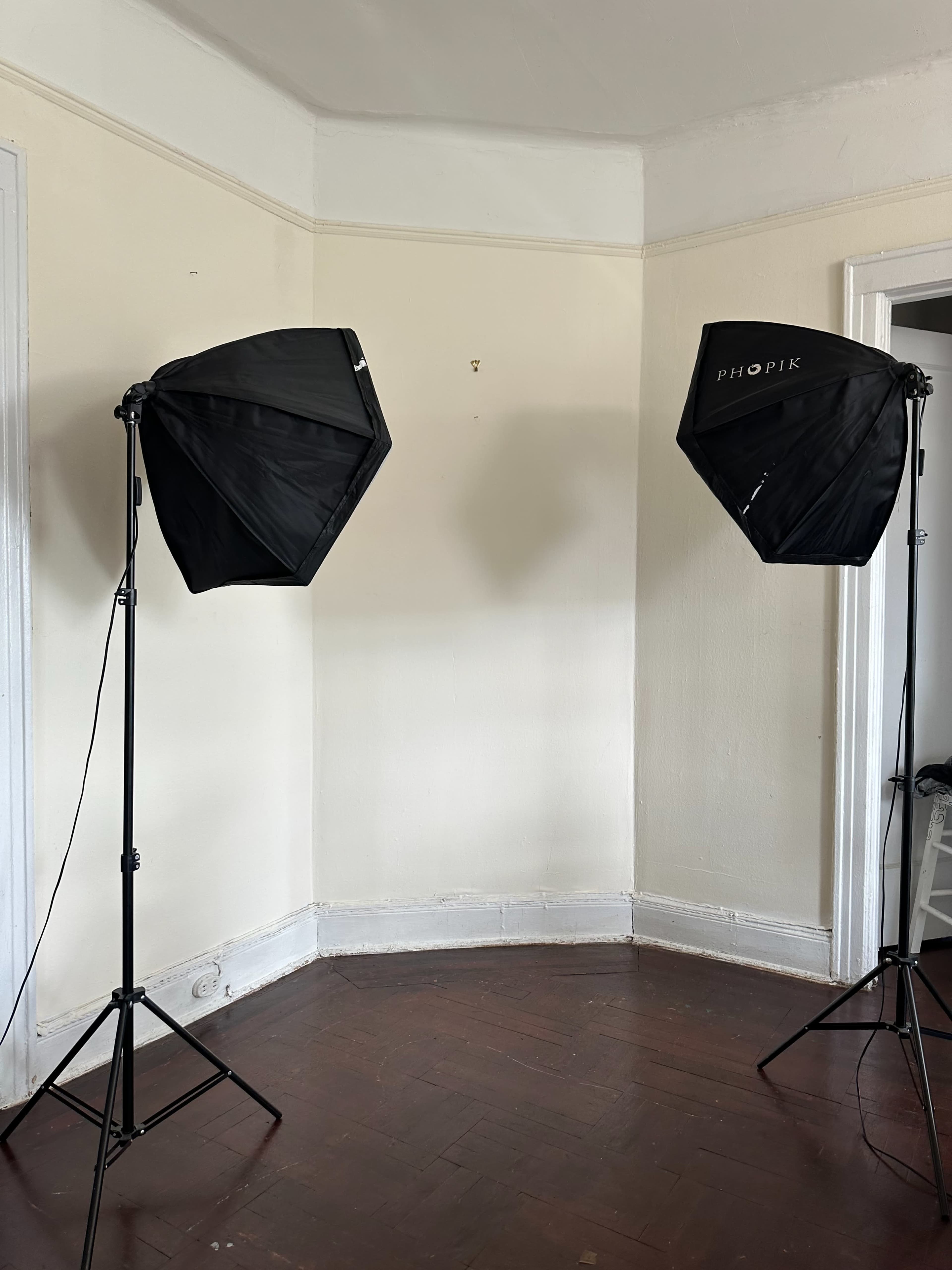 The image shows two black softbox lights set up on stands in a corner of a room with hardwood floors and plain walls.