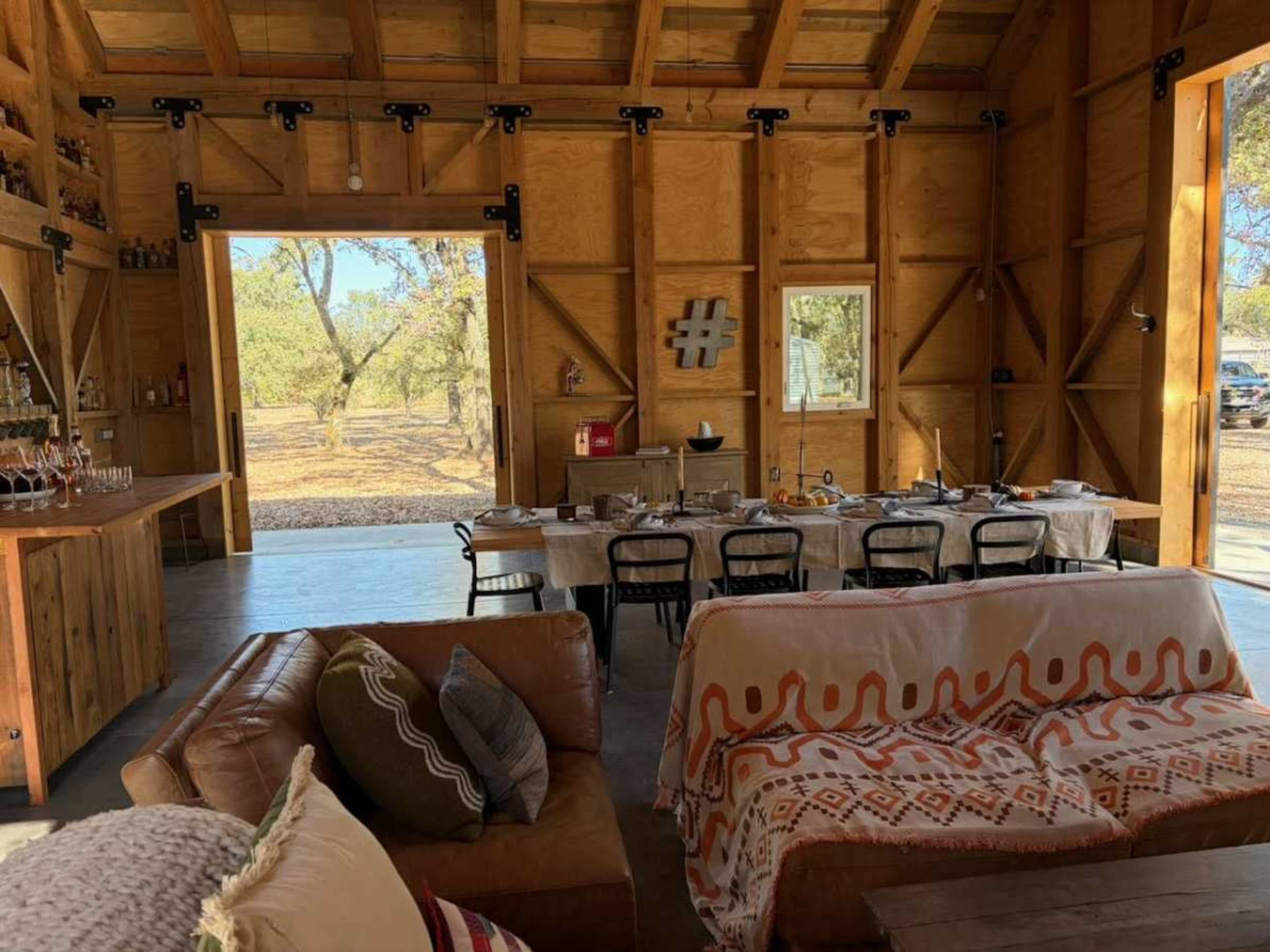 The interior of a wooden barn features a dining table set for a meal, a seating area with a couch, and large open doors that display a view of the outdoors.