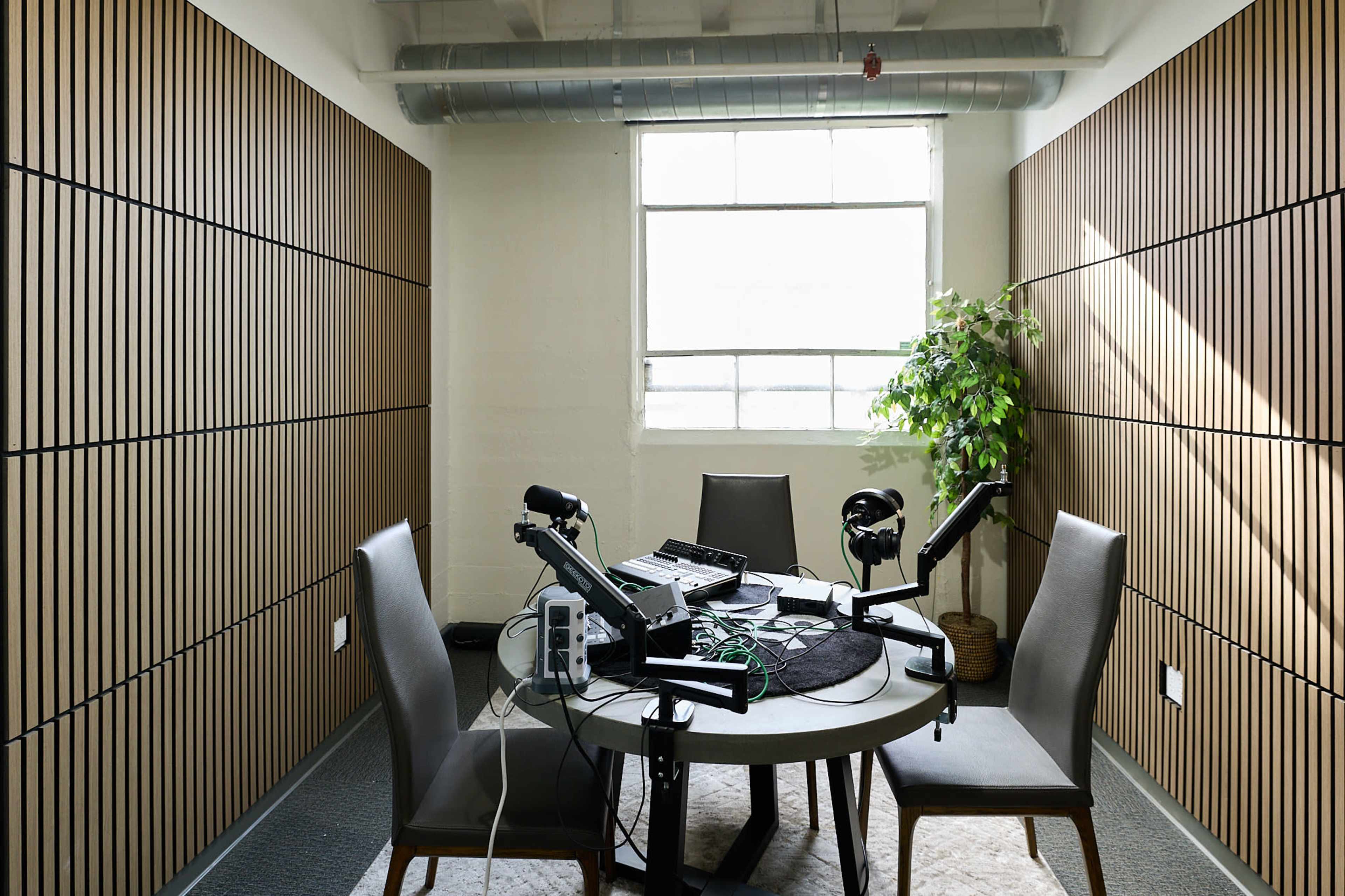 The image shows a small recording studio with a round table, two chairs, microphones, and audio equipment, featuring soundproofed walls and a window.