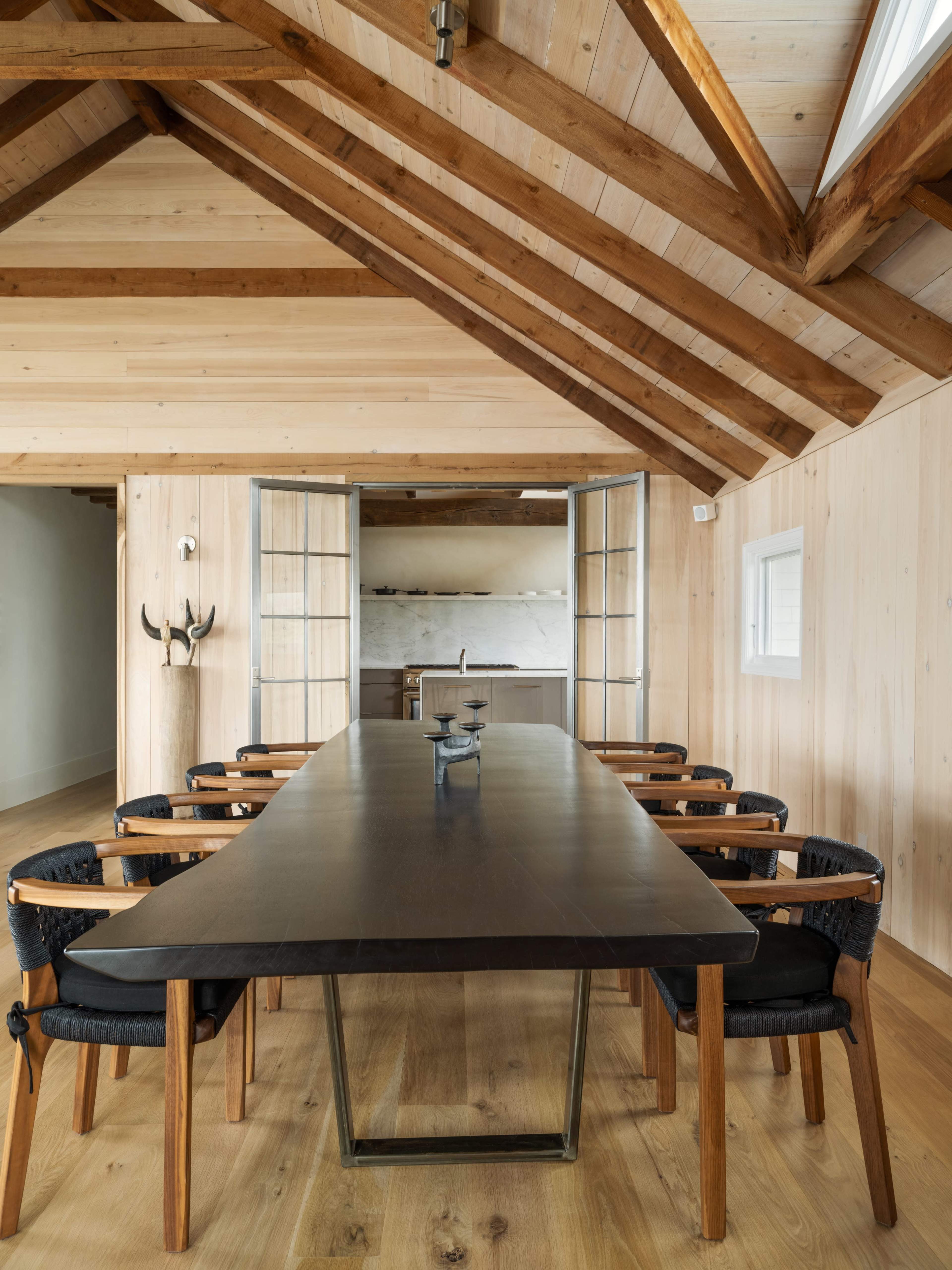 A spacious dining room features a long wooden table surrounded by black-upholstered chairs, with a vaulted wooden ceiling and large windows.