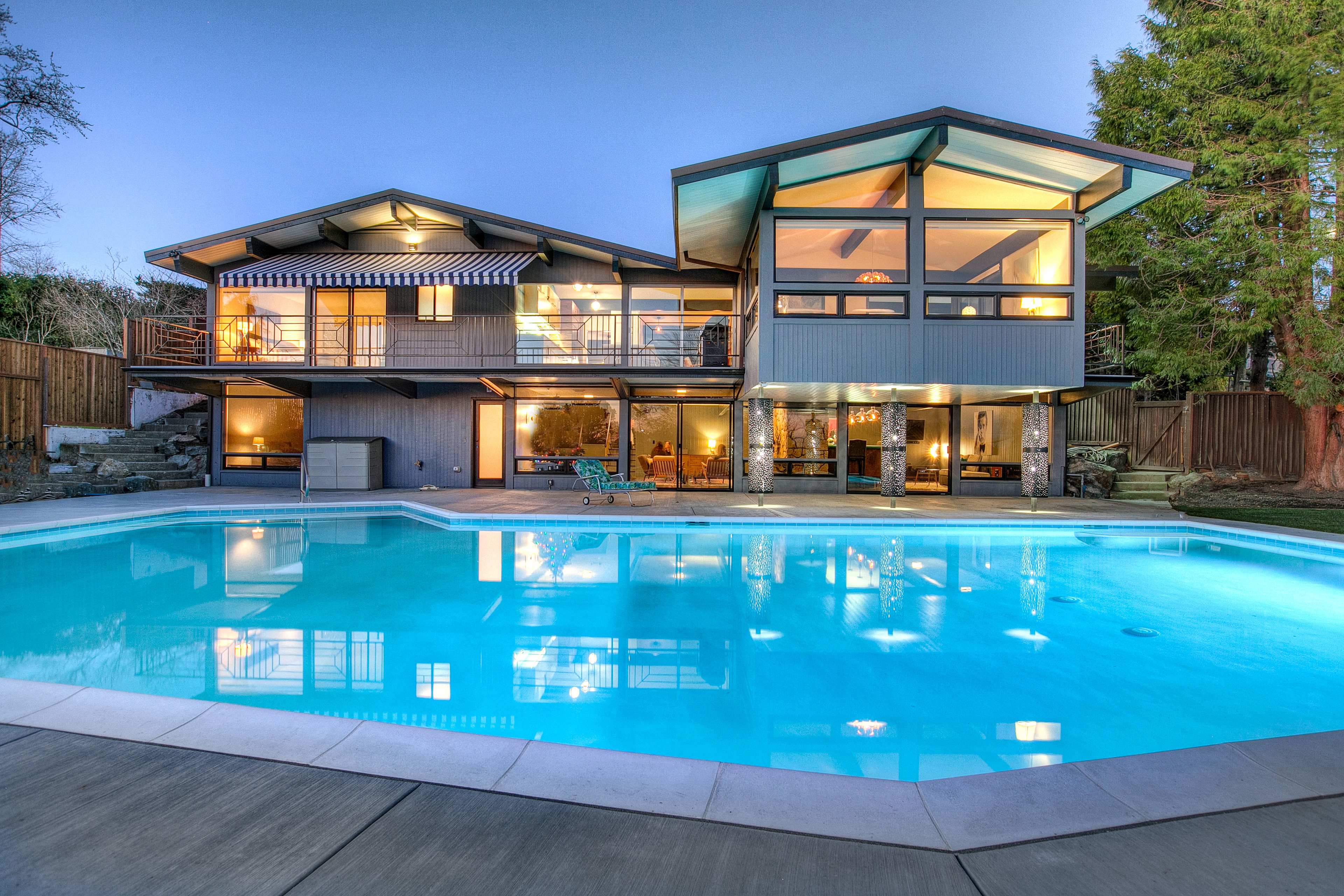 A modern two-story house with large windows, overlooking a rectangular swimming pool.