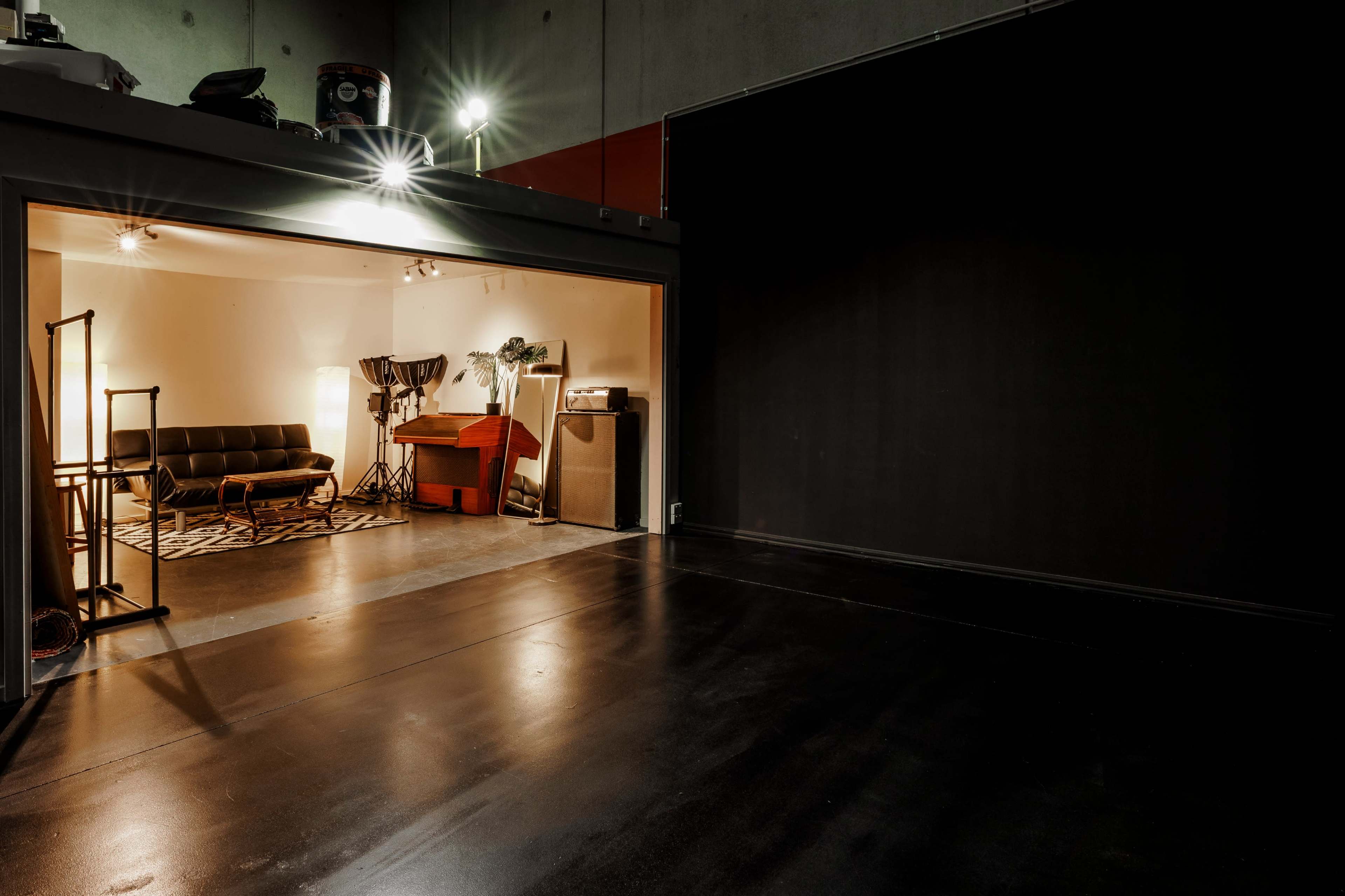 The image shows a spacious studio with a well-lit area containing a sofa, a piano, and various lighting equipment, alongside a large dark wall opposite the entrance.