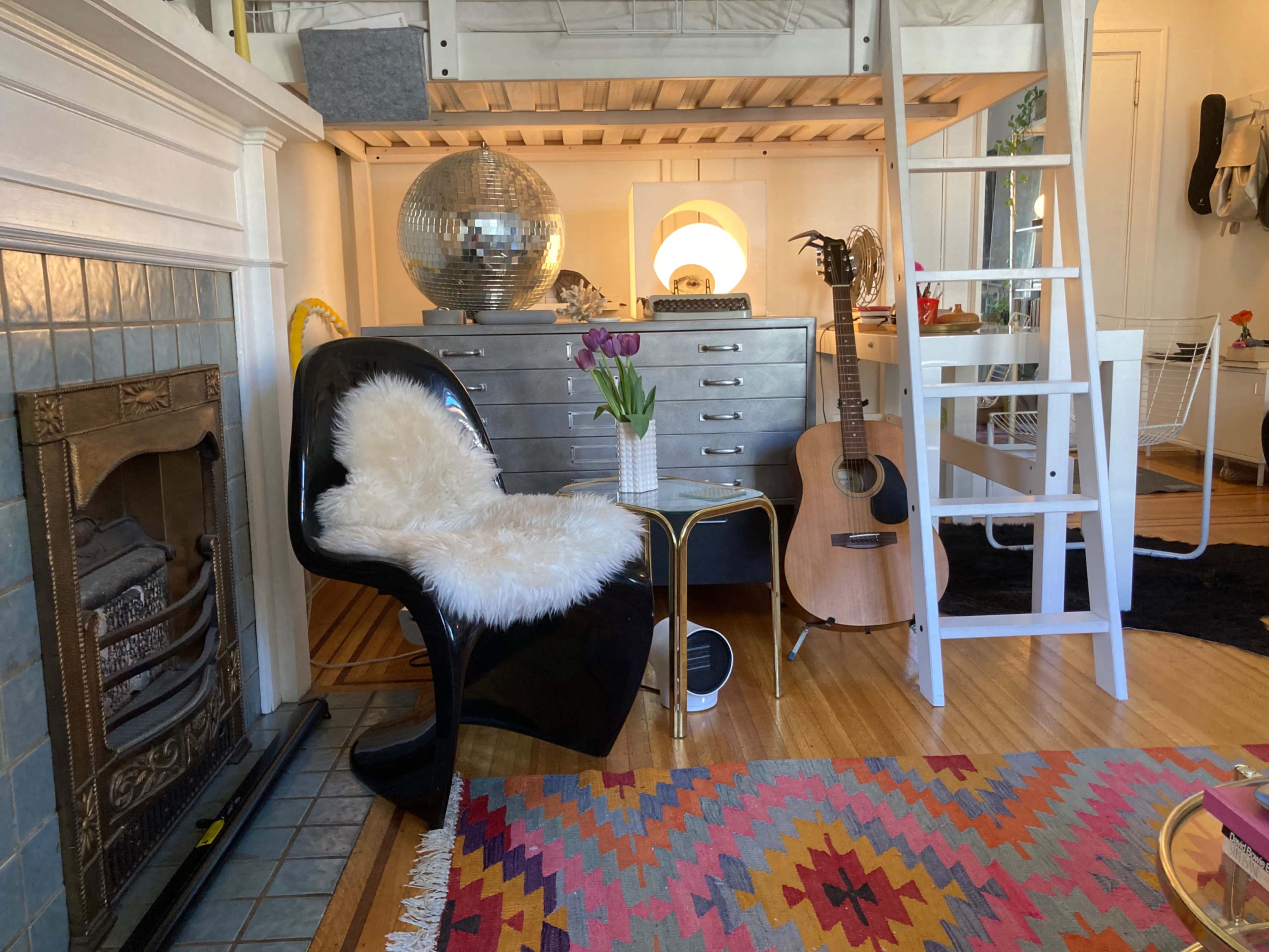 The image shows a cozy interior featuring a black chair with a white fur throw, a wooden ladder leading to a lofted area, a silver dresser, a guitar, and decorative elements like a disco ball and fresh flowers.