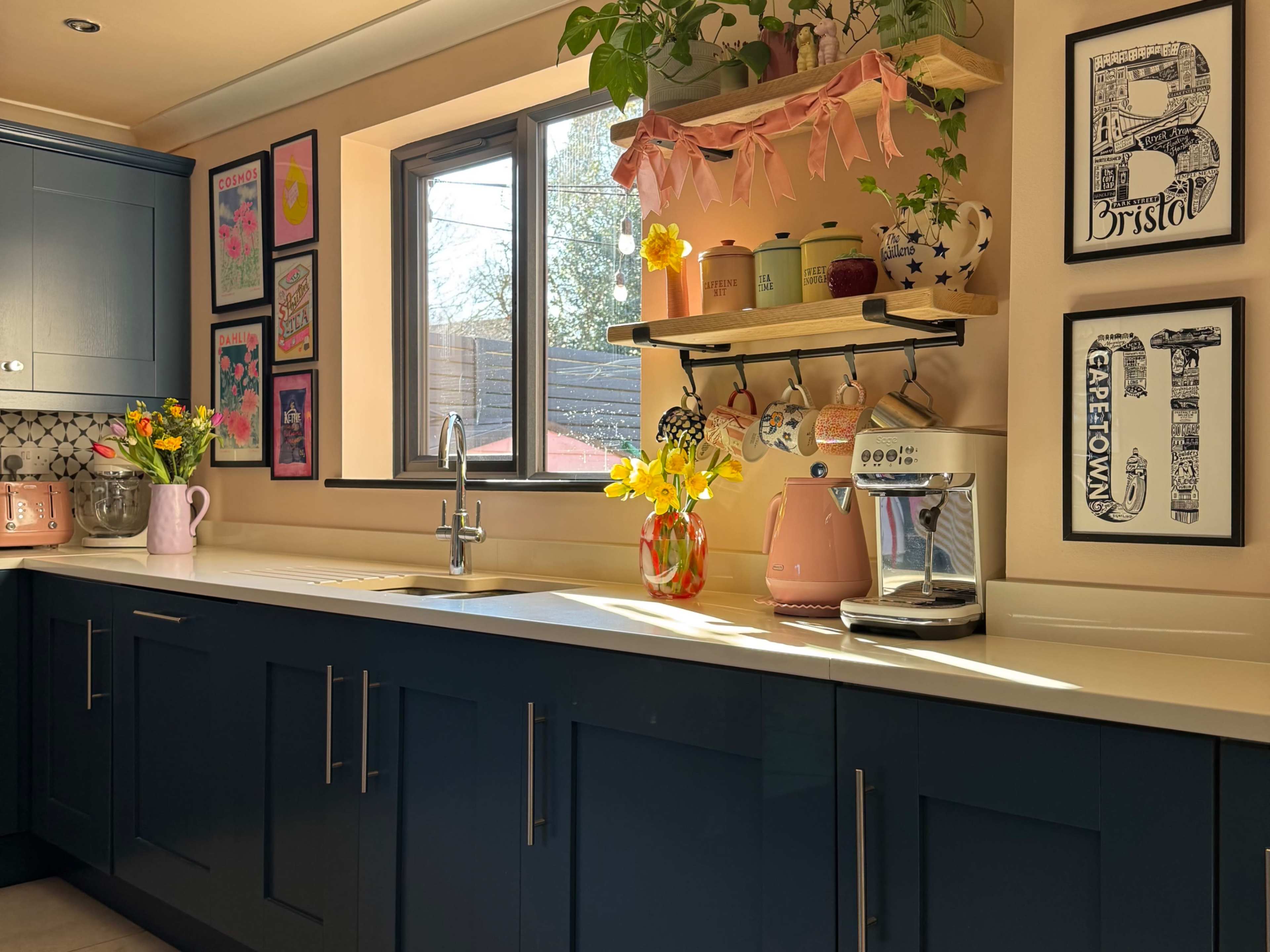 A modern kitchen features navy blue cabinetry, a white countertop, and a window adorned with potted plants and decorative items, illuminated by natural light.