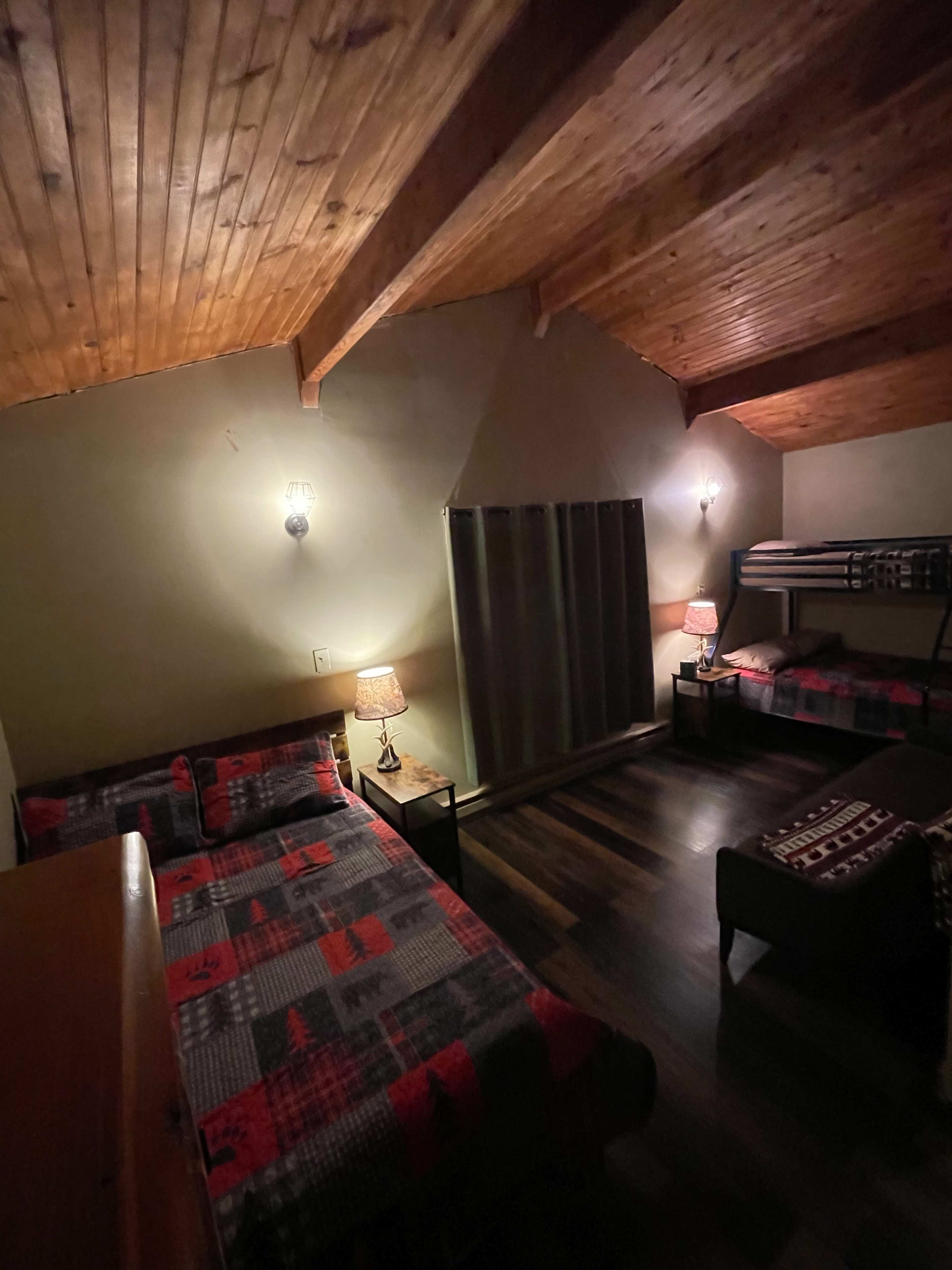 The image shows a dimly lit bedroom with two beds and a bunk bed, featuring wooden beams on the ceiling and a large window covered by curtains.