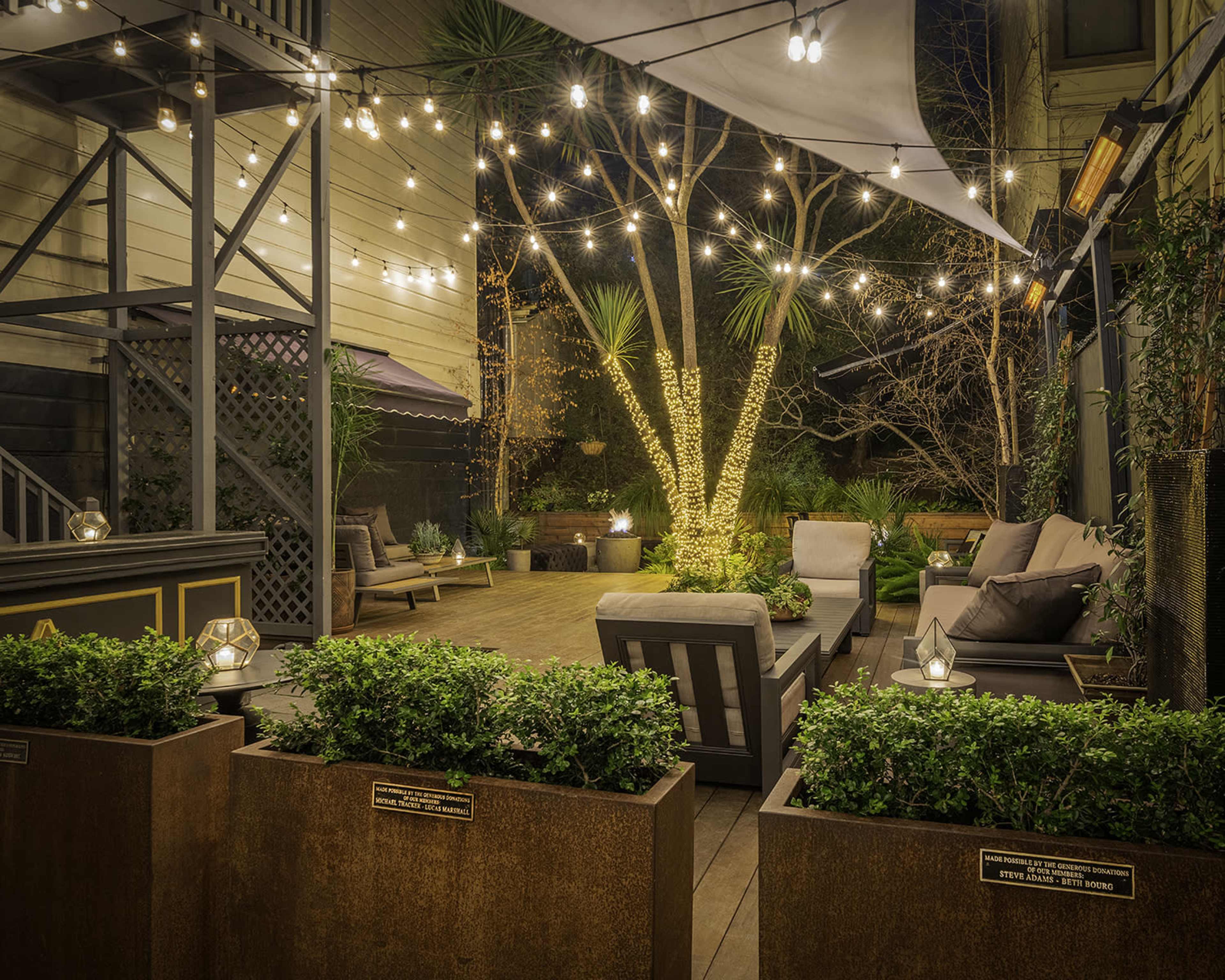 The image shows a beautifully lit outdoor patio area featuring comfortable seating, potted plants, and string lights draping overhead.